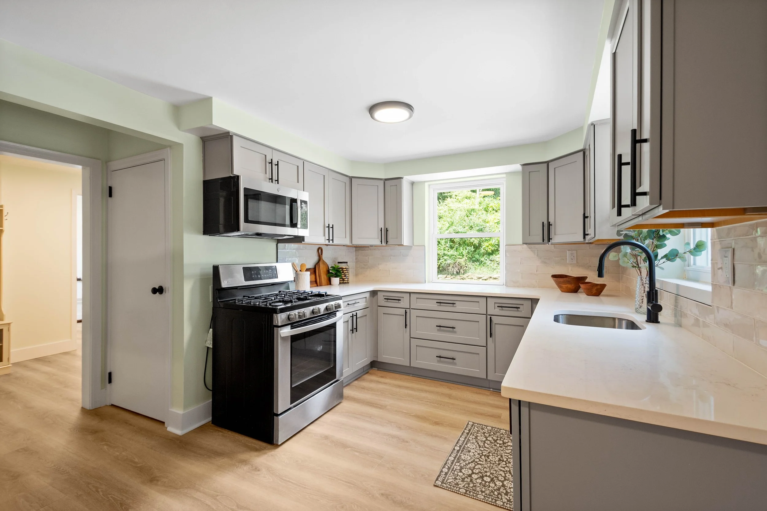 A modern kitchen with light green walls, white cabinets, black hardware, a stainless steel oven and microwave, a window overlooking greenery, a black faucet over a sink, and wooden bowls on the countertop.