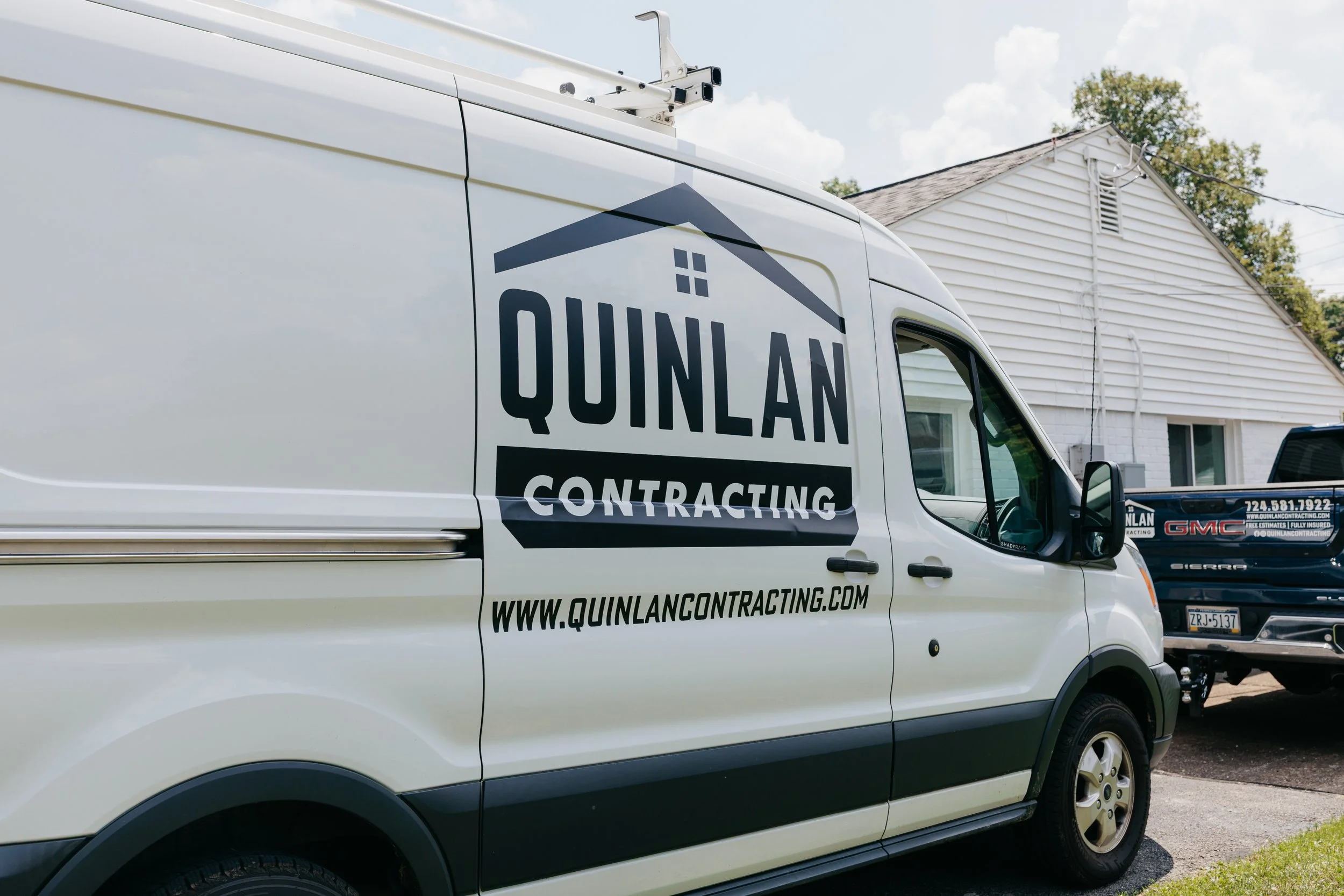 A white contracting van with the logo 'Quinlan Contracting' and website 'www.quinlancontracting.com' is parked outdoors near a house. Another truck is visible in the background.