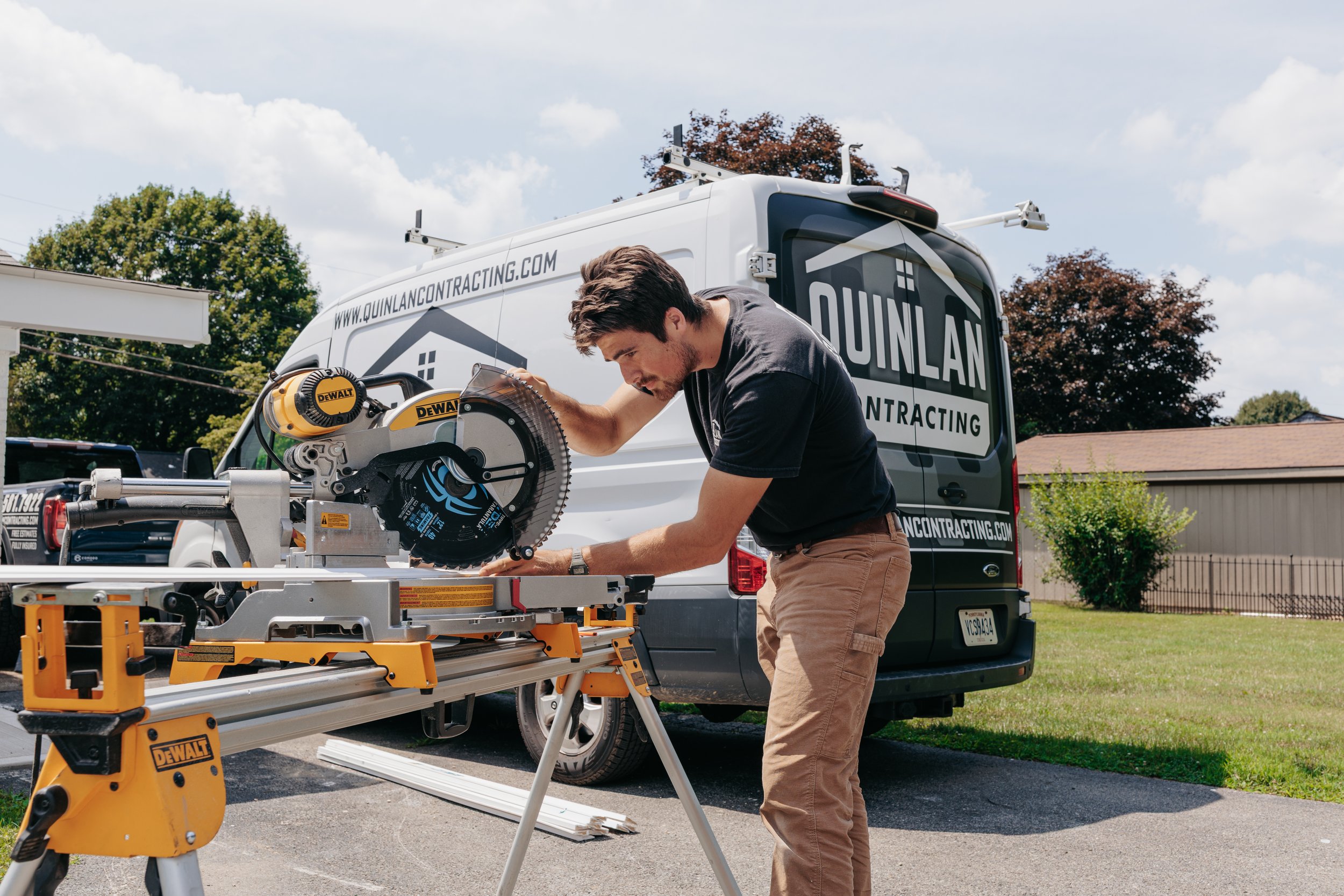 A man working with a circular saw on a workbench in front of a white van with 'Quinlan Contracting' branding, outdoors on a residential street with trees and a blue sky.