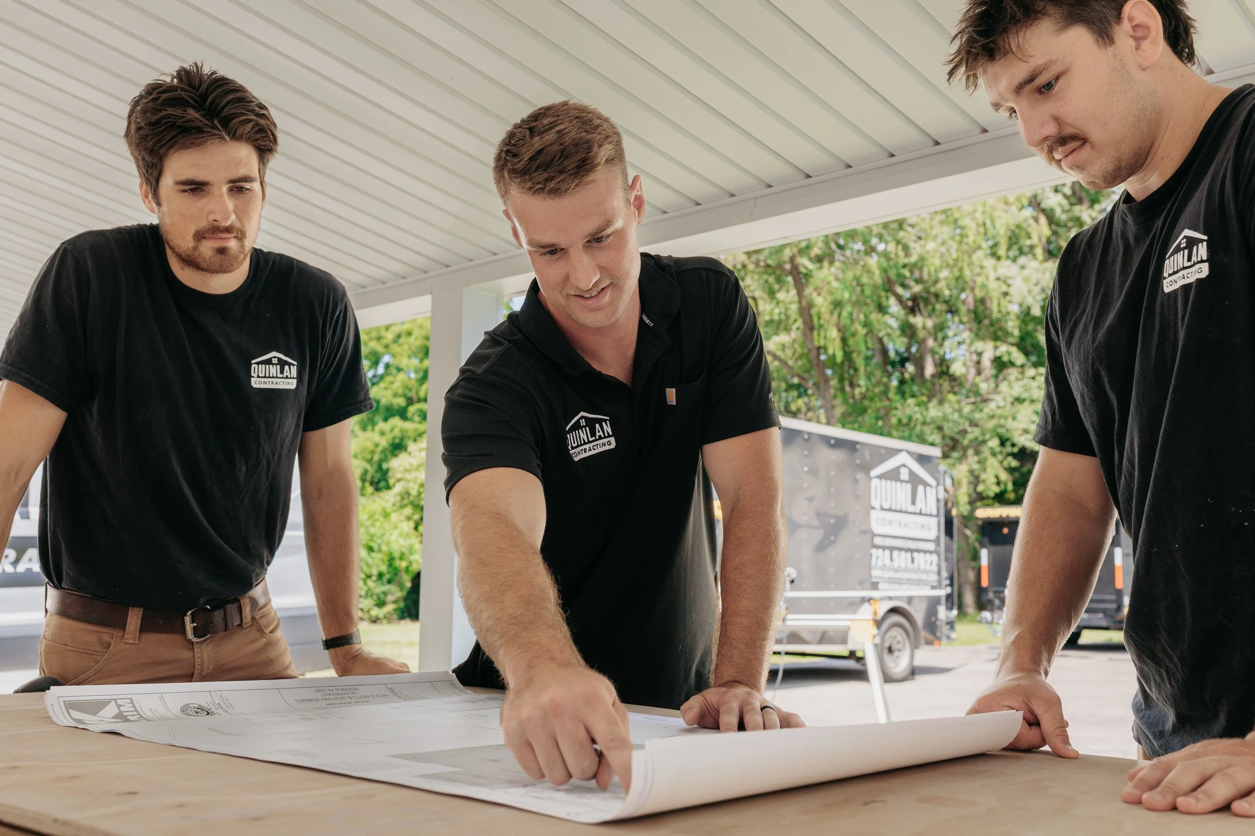 Three men reviewing blueprints at an outdoor construction site, wearing black shirts with 'Quinlan Contracting' logos.