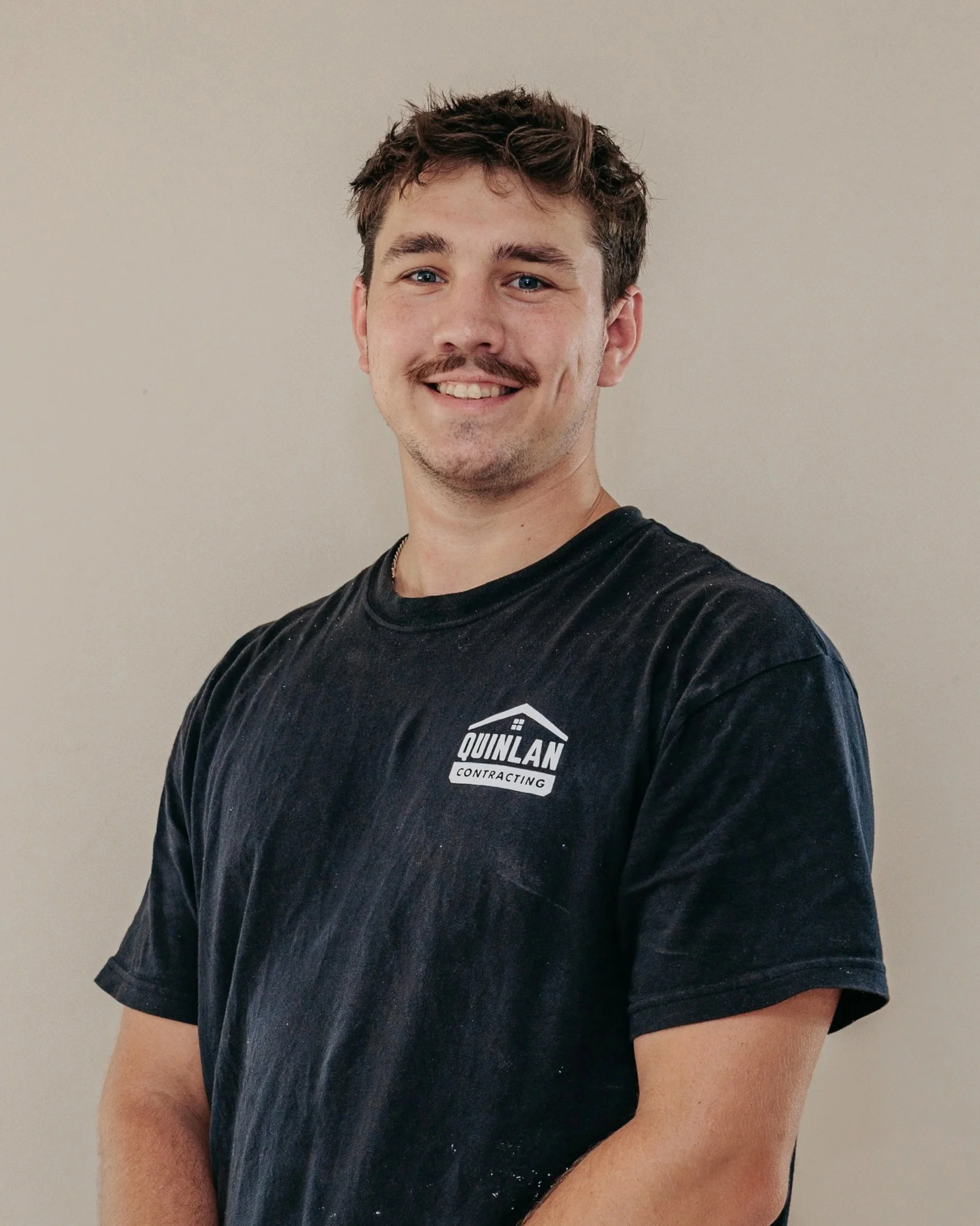 A young man with light brown, slightly curly hair, blue eyes, and a mustache, smiling while wearing a black t-shirt with a company logo, standing against a plain, light-colored background.
