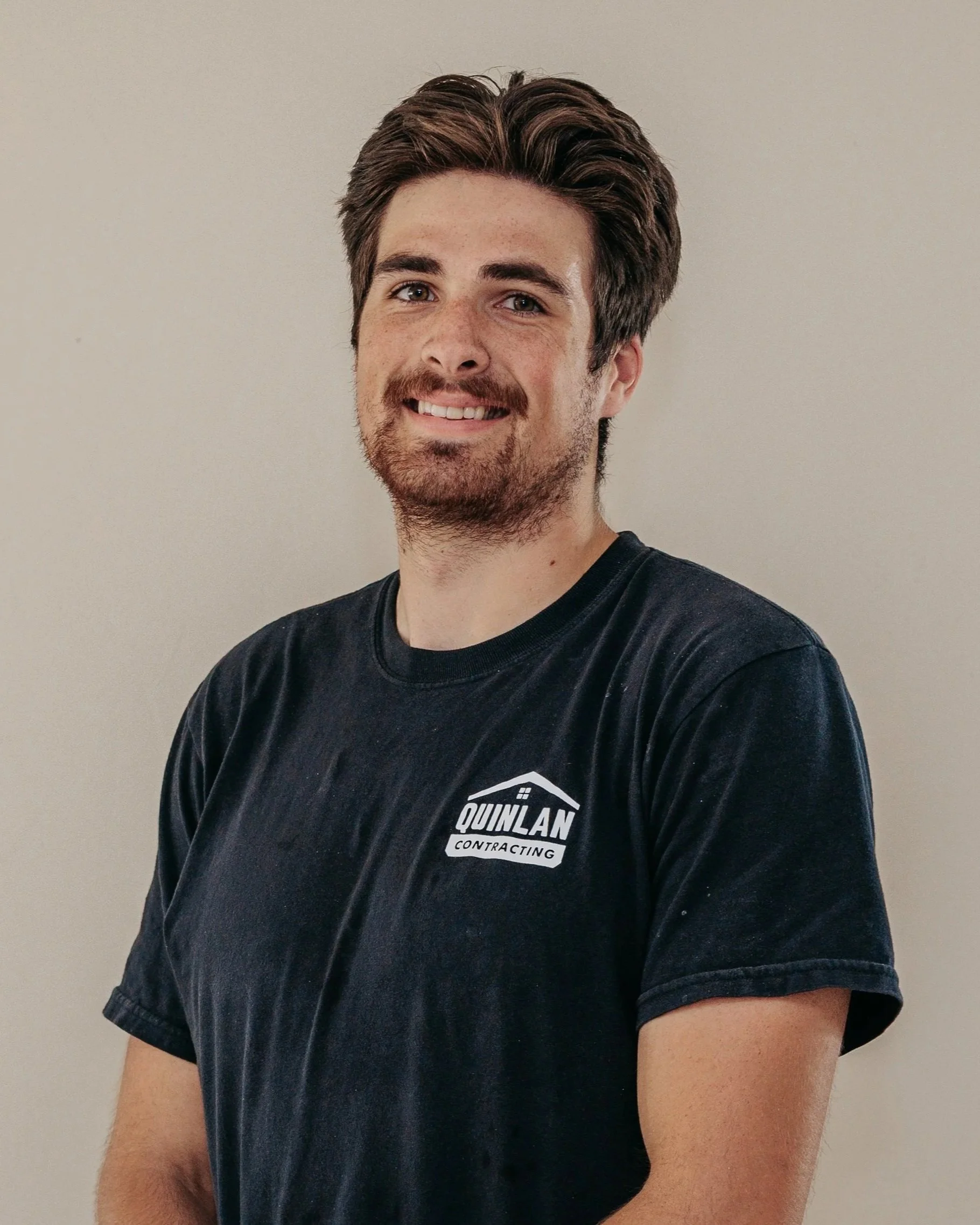 A young man with brown hair and a beard wearing a black t-shirt with a logo that reads 'QUINLAN CONTRACTING' standing against a plain light-colored wall.