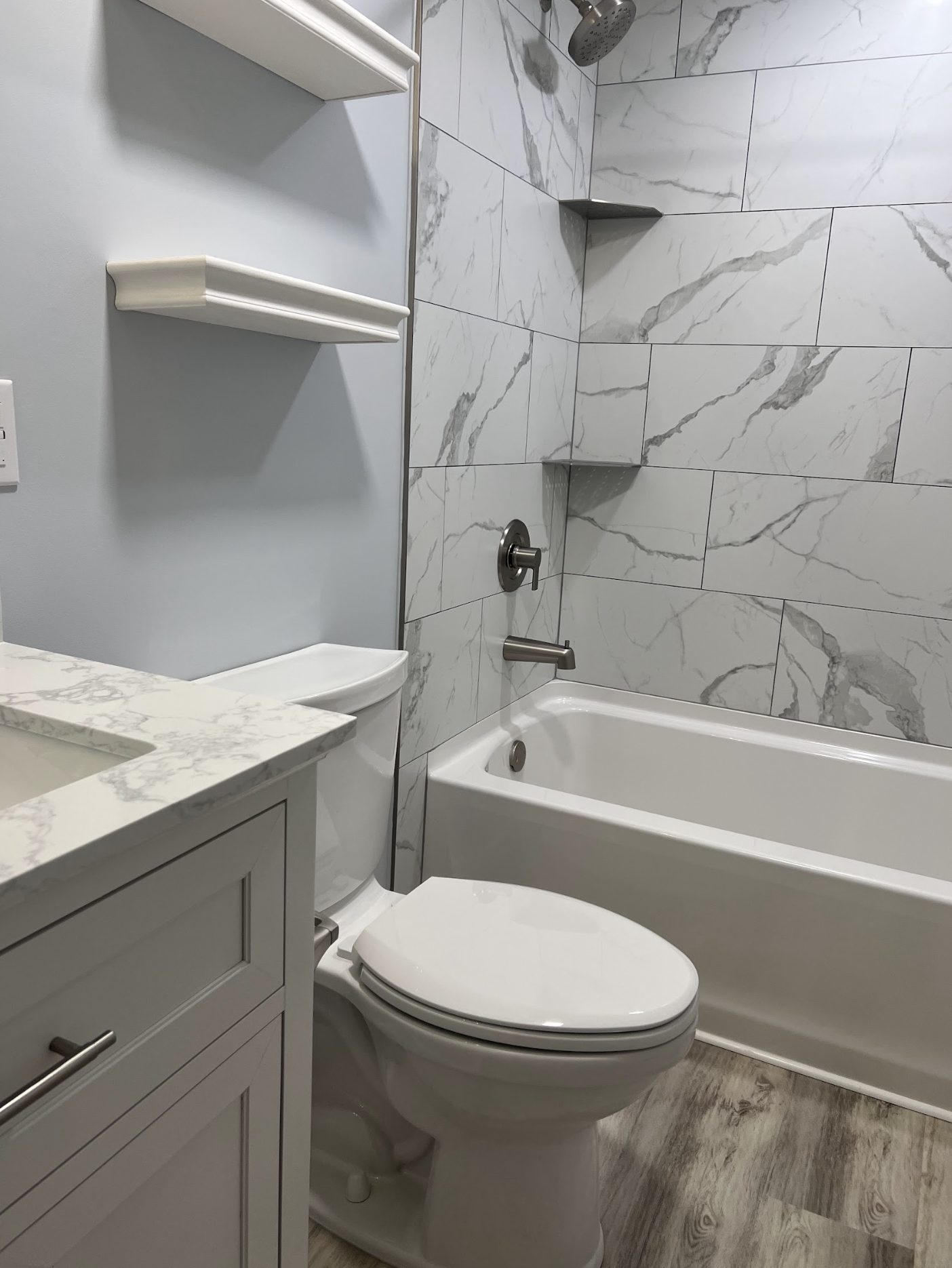 A bathroom with a white toilet, a white vanity with a marble countertop, and a bathtub with a marble tile surround. There are two corner shelves in the shower area and a showerhead above the bathtub.