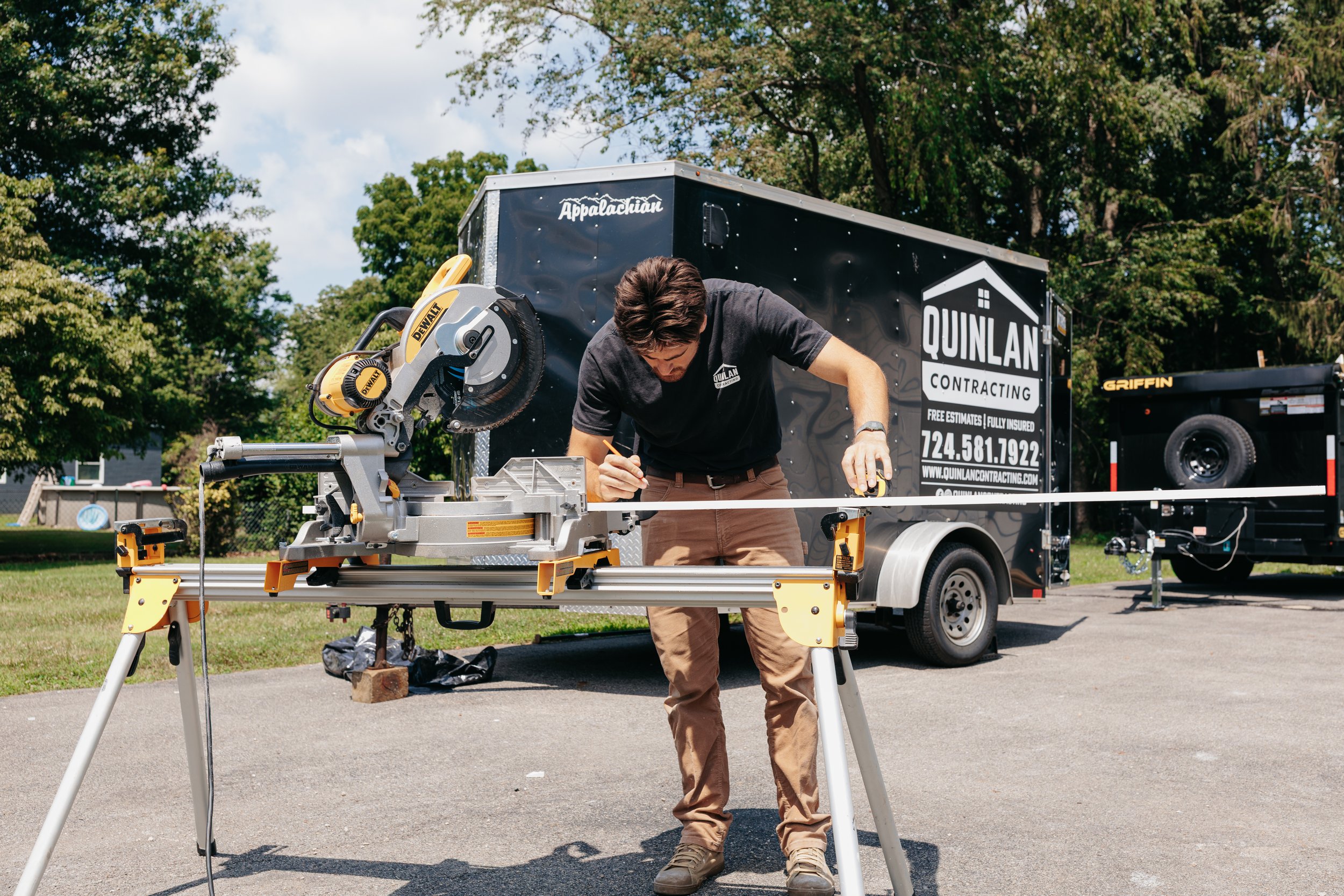 A man working on a large sliding miter saw setup outdoors, with a black trailer in the background labeled 'Quinlan Contracting' and a yellow DeWalt logo on the saw. The scene is in a suburban area with trees and houses visible.