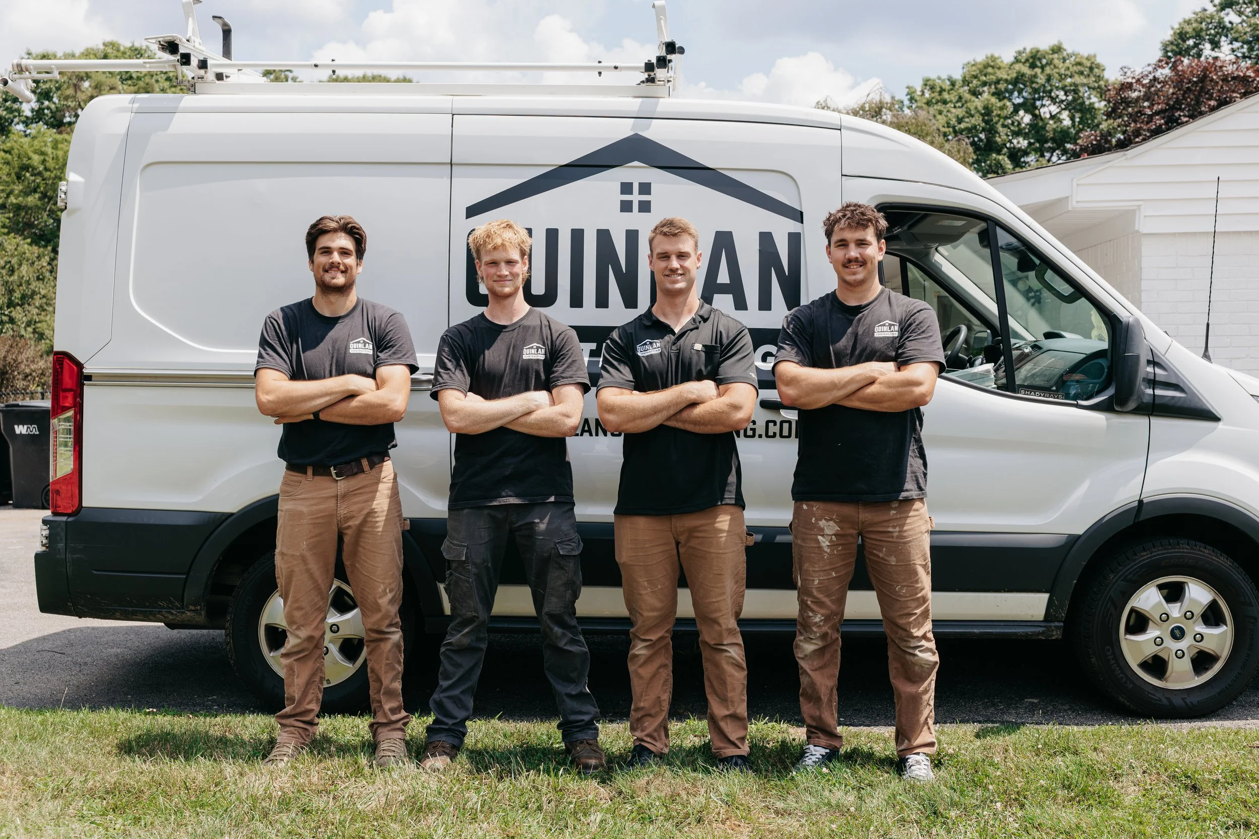 Four young men standing with arms crossed in front of a white work van with a company logo that reads "QUINLAN" and a graphic of a house roof on the side. They are outdoors in front of a house, with trees and a partly cloudy sky in the background, wearing work shirts with the company logo.