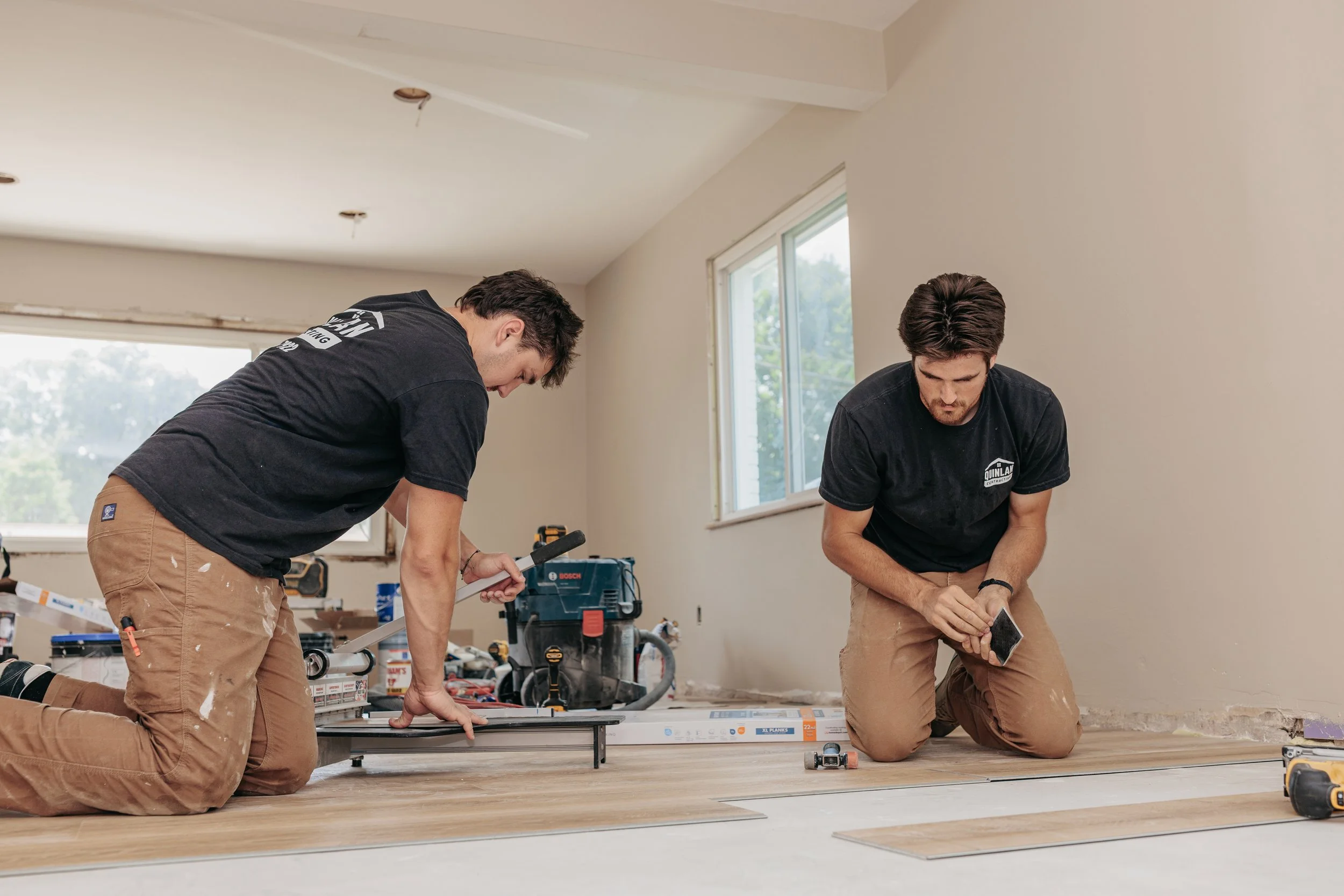 Two men kneeling on a wooden floor installing flooring in a room with large windows, surrounded by tools and construction supplies.