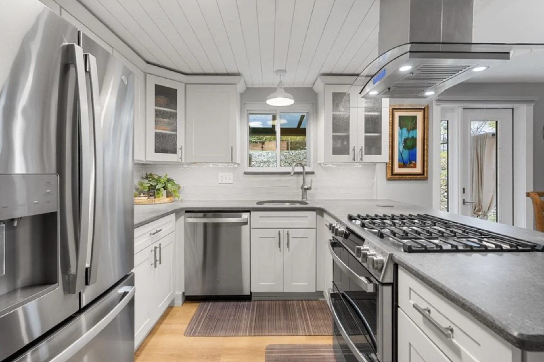 Modern kitchen with white cabinets, stainless steel appliances, a gas stove, a small window above the sink, and artwork on the wall. There is a small plant on the counter and a brown rug on the wooden floor.