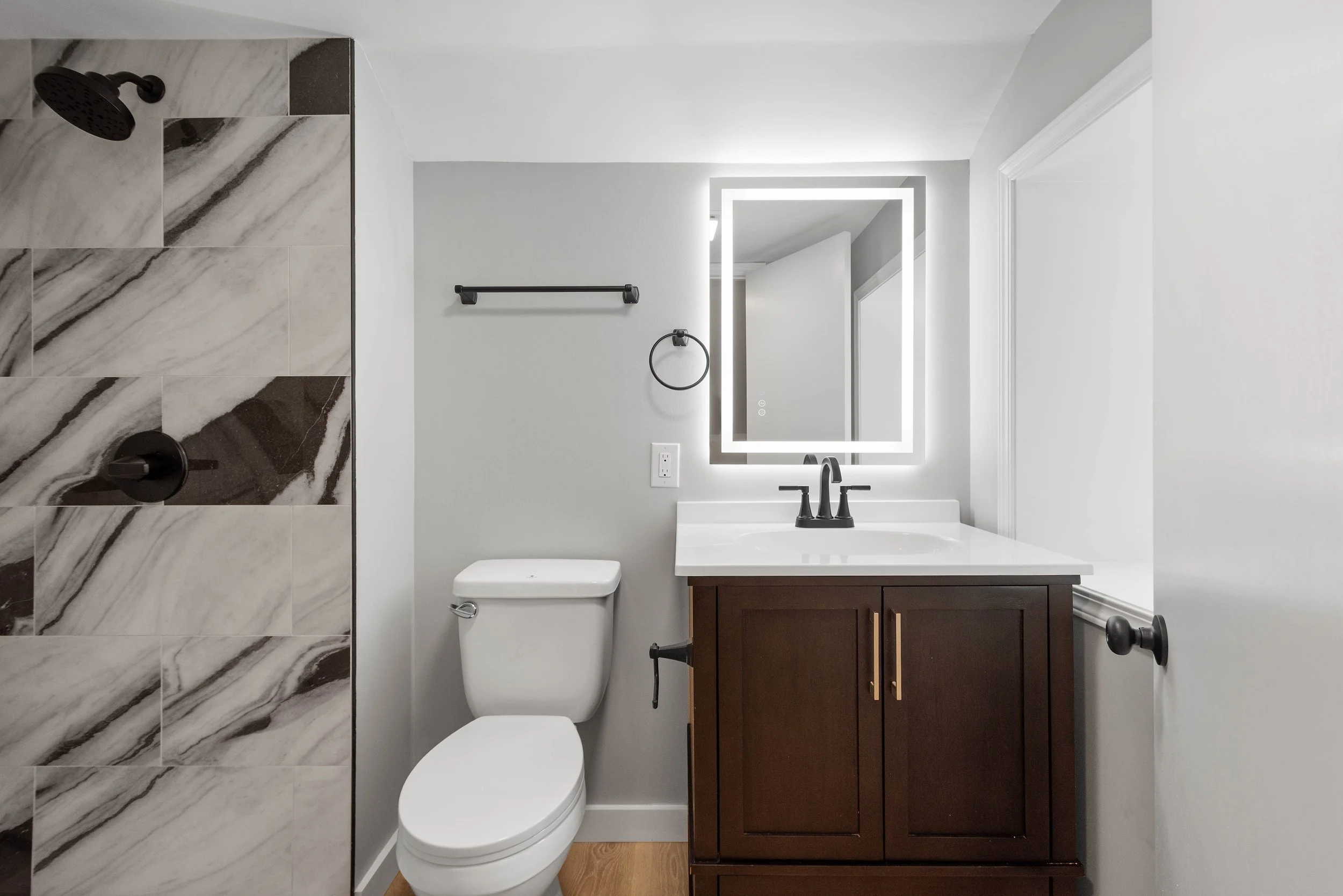 A modern bathroom with a shower featuring marble tiles, a white toilet, a dark wood vanity with a white countertop, and a backlit rectangular mirror.