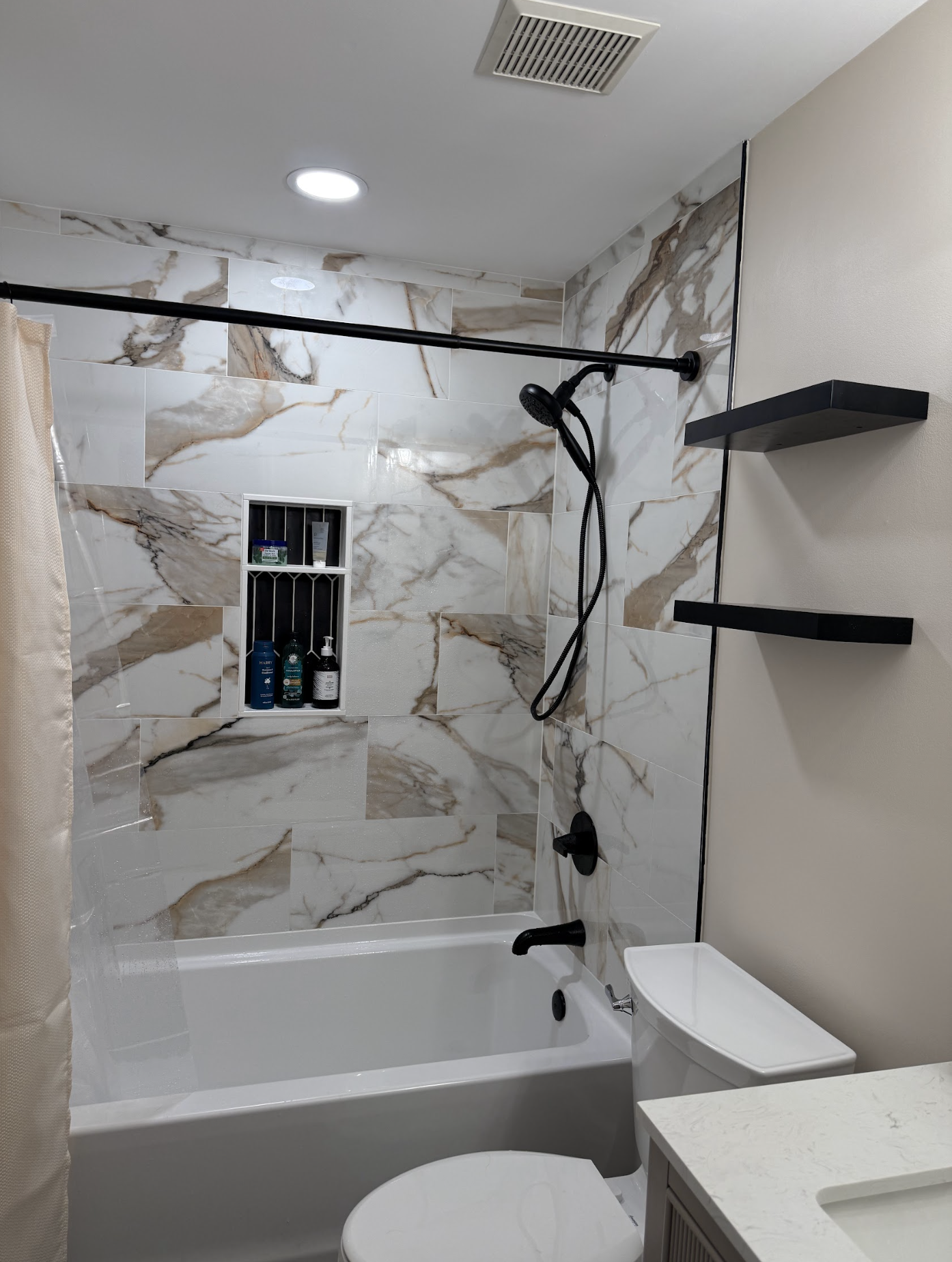 Bathroom with marble tile walls, bathtub, shower head, black fixtures, built-in shelves with toiletries, toilet, and beige curtain.