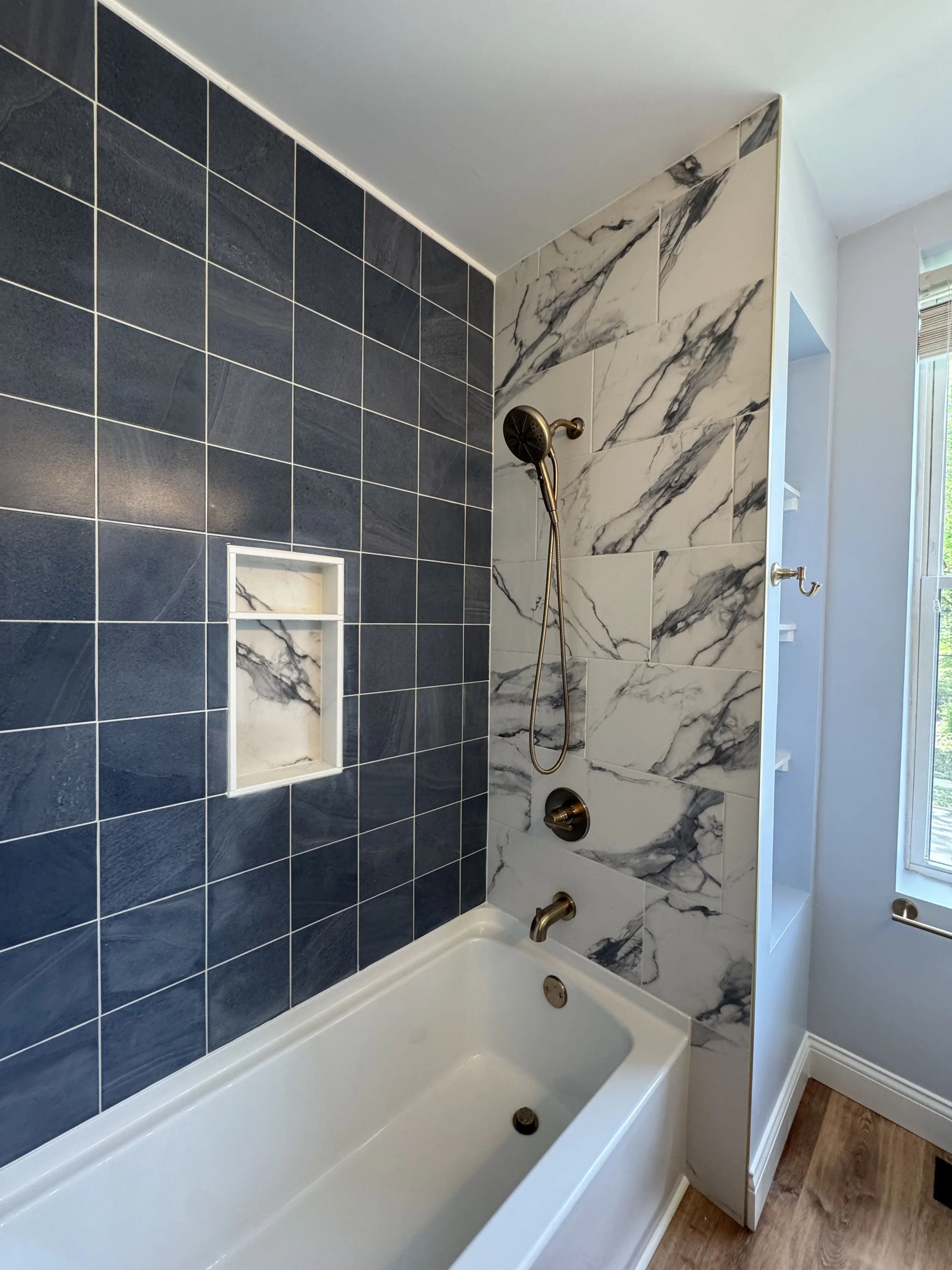 Bathroom with a bathtub, a shower head with a brass finish, dark blue wall tiles, and white marble tiles with gray veining on the wall, with a built-in niche in the tile wall.