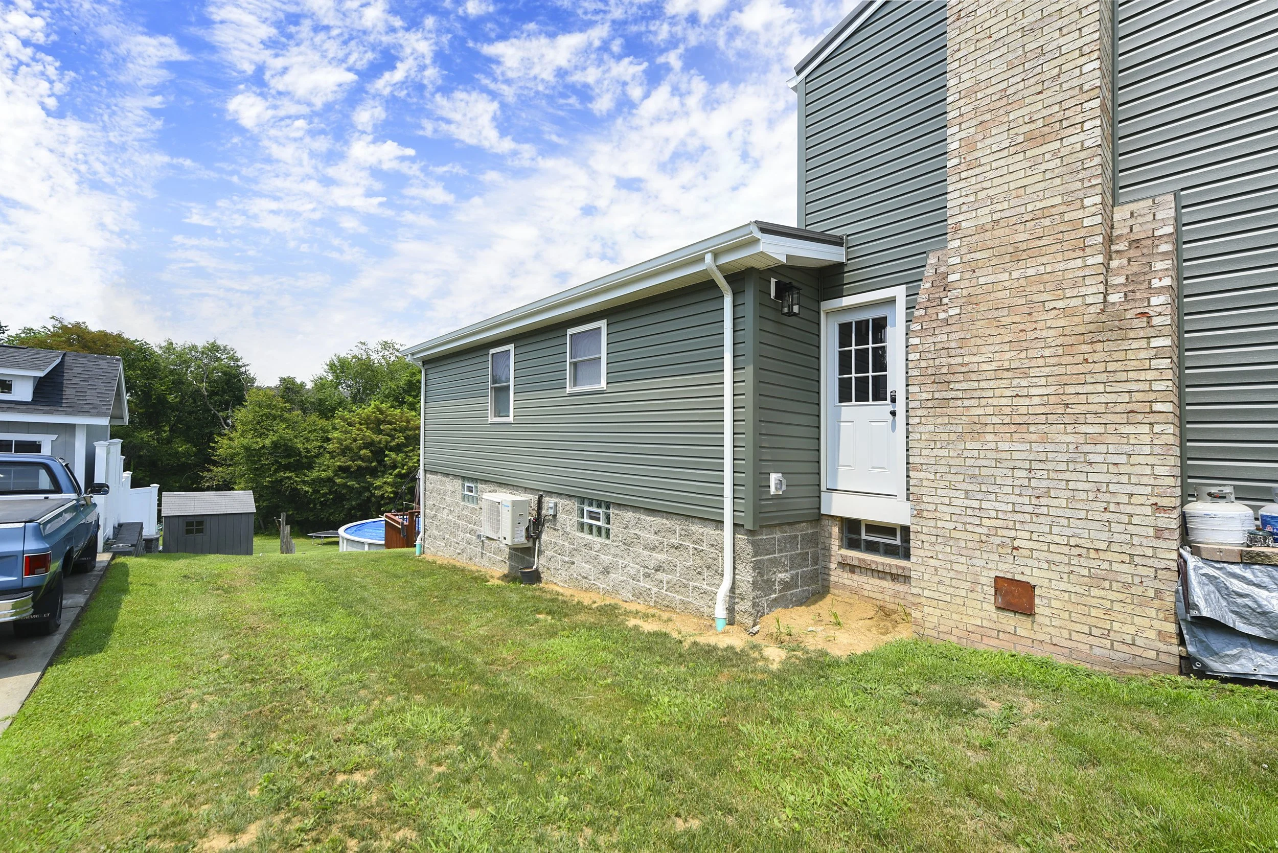 Backyard view of a house with green siding, brick chimney, and a white door, with a grassy lawn and a blue sky with clouds.
