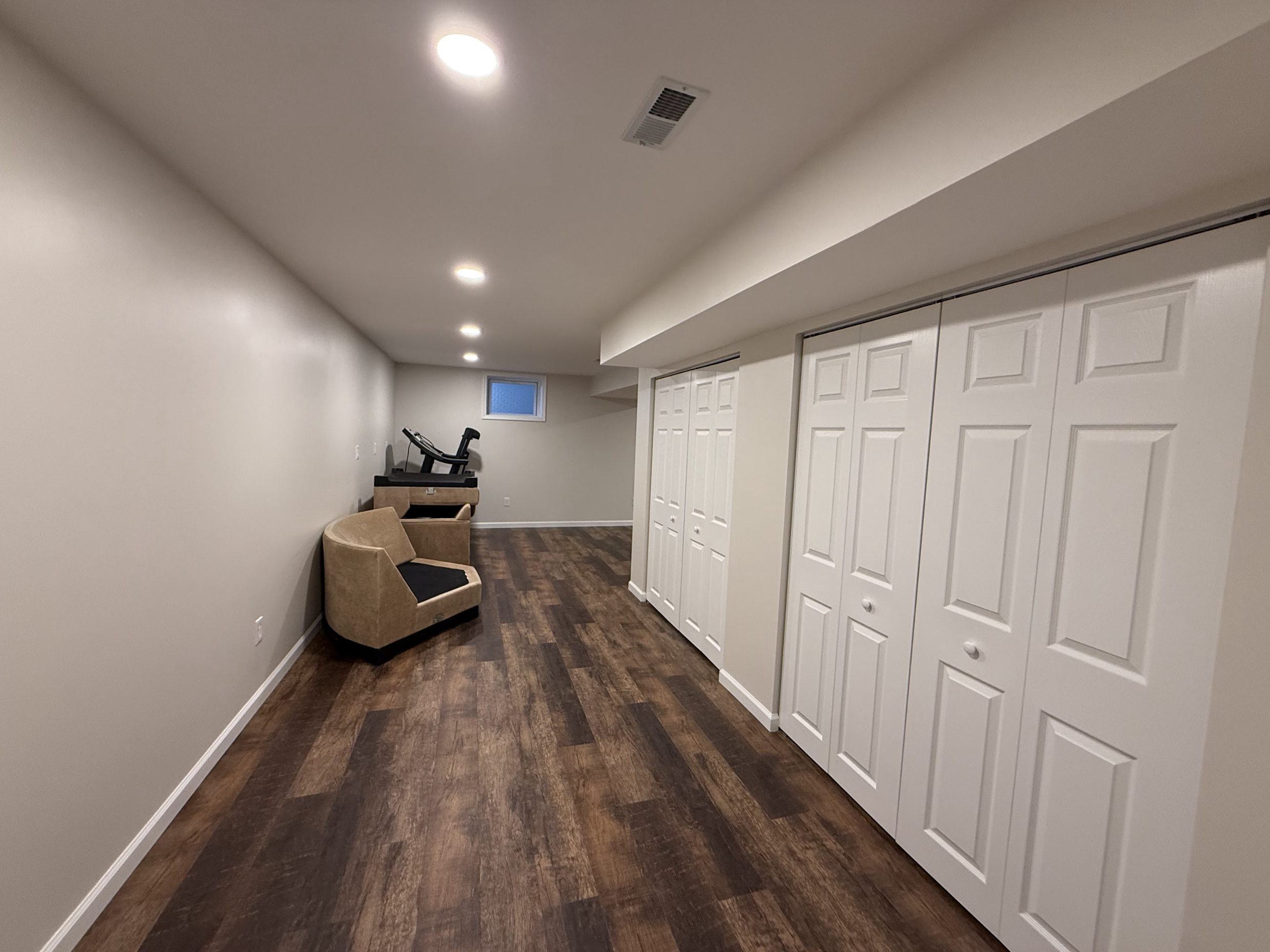 A basement room with beige walls, dark hardwood flooring, white double closet doors, recessed lighting, a small window, and sports equipment including a treadmill and exercise bike.
