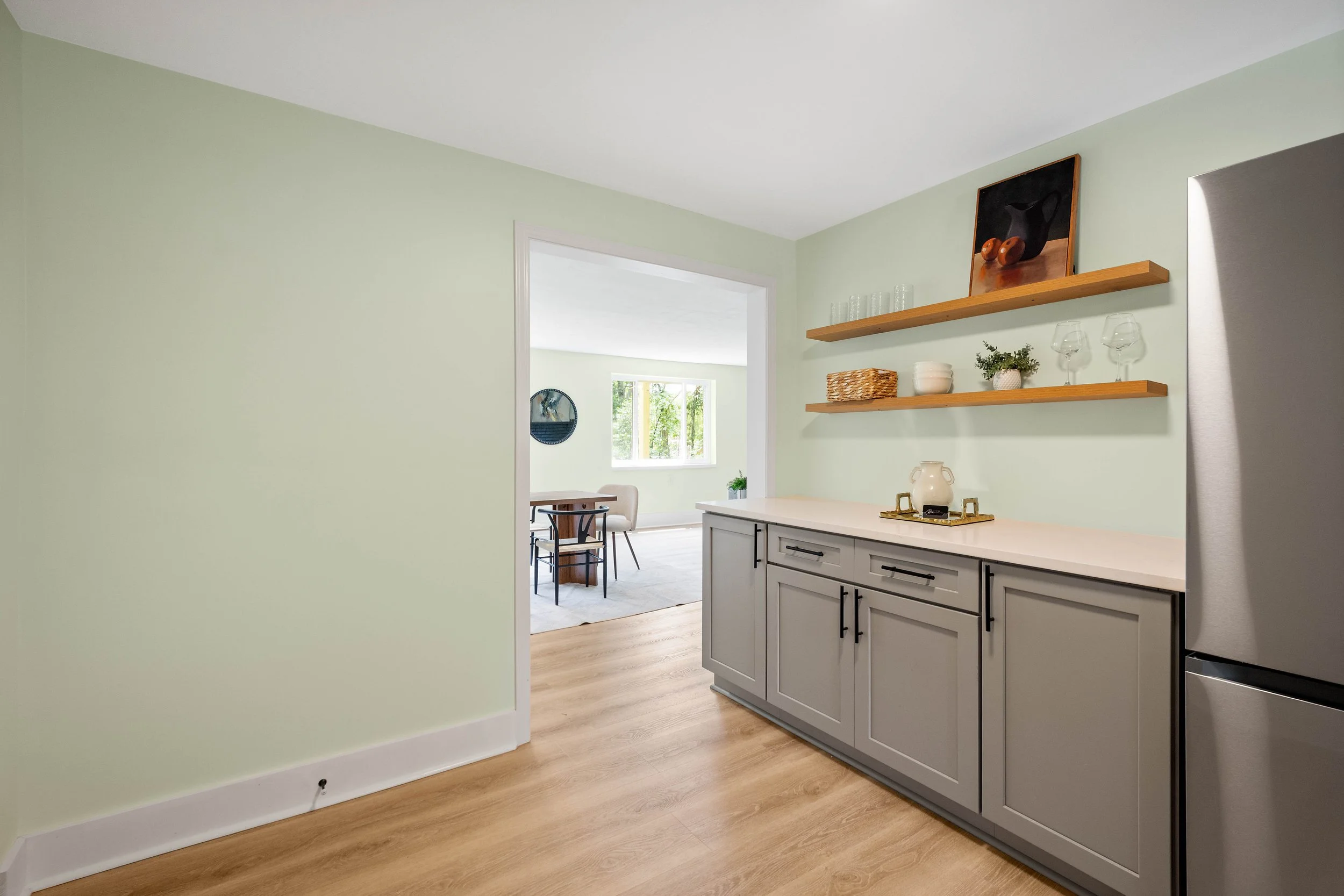 Kitchen with light green walls, gray cabinets, wooden floating shelves, and a view into a dining area with a window and some chairs.