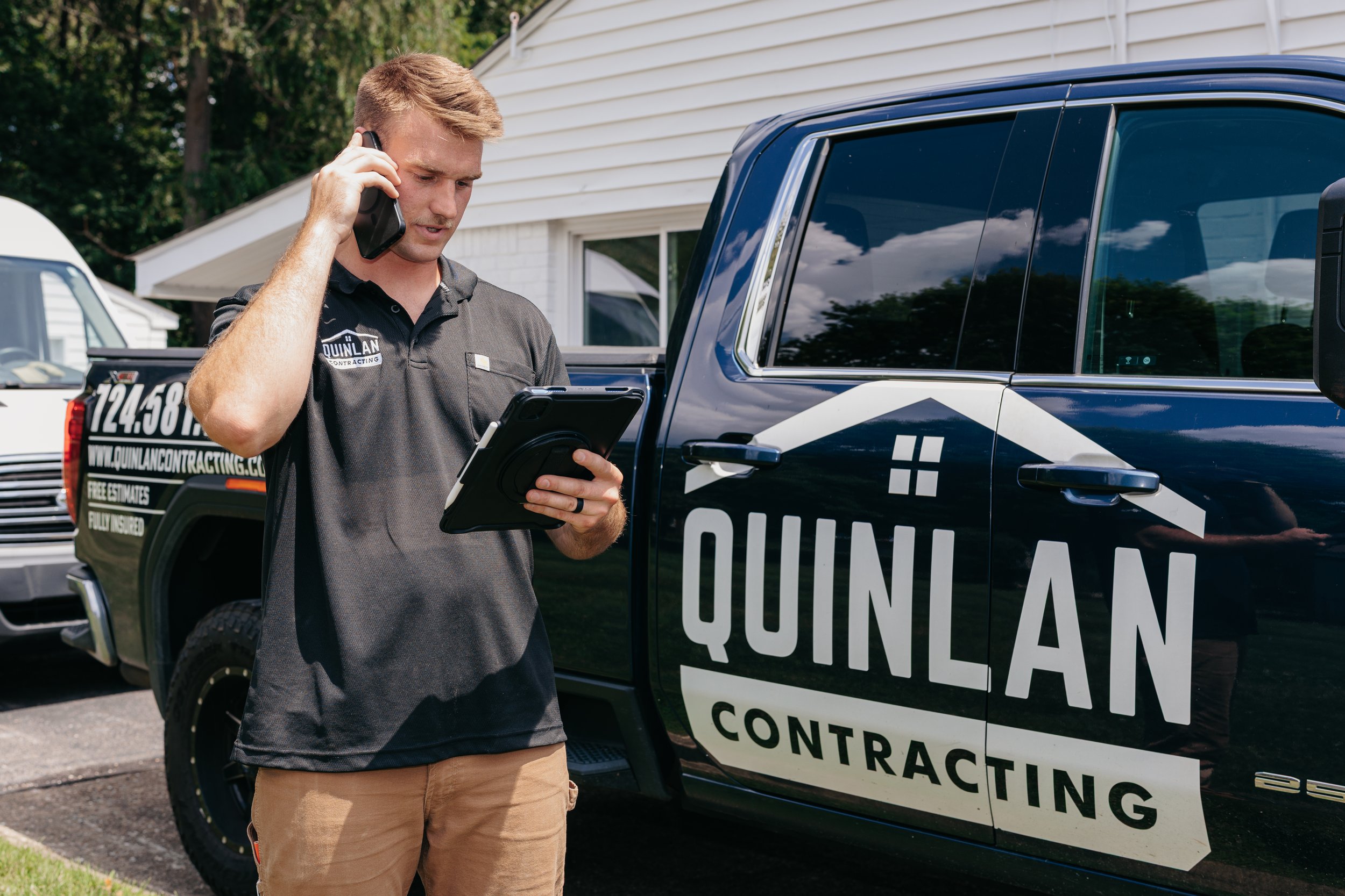 A man with blonde hair wearing a black polo shirt with a logo, standing beside a black and white contracting vehicle, on his phone and holding a tablet.