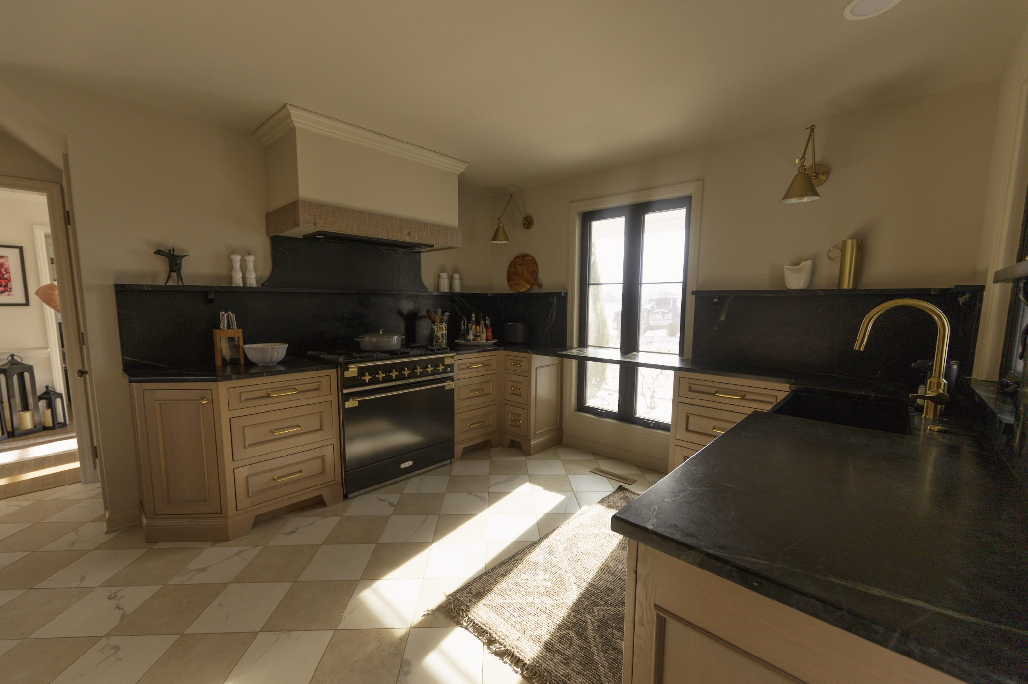 Kitchen with beige cabinets, black countertops, a black stove, and large windows, lit by gold wall-mounted lights.