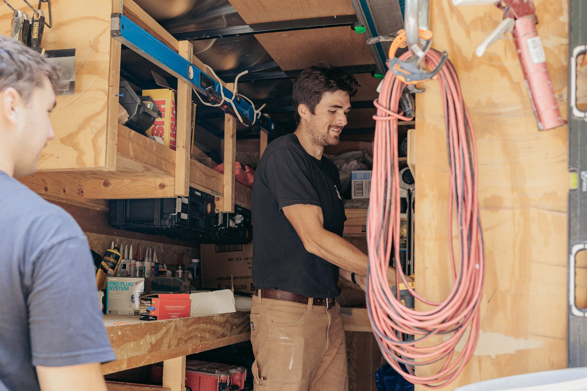 Two men working in a workshop or garage, with one man smiling and working on electrical wiring or equipment, and the other man partially visible in the foreground.