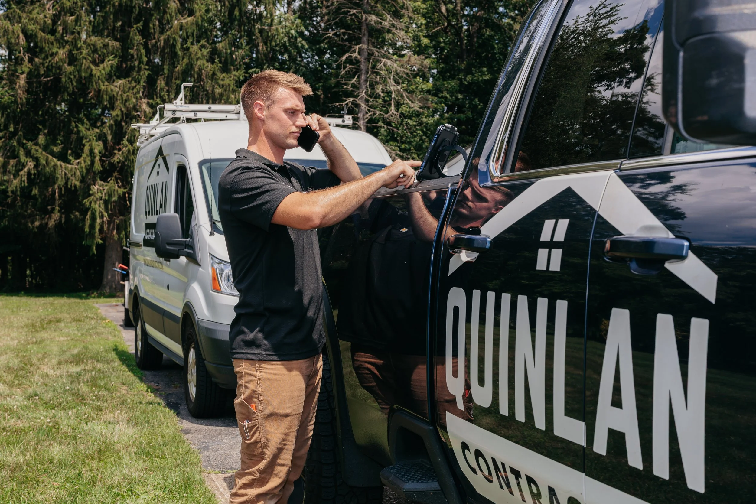 A man with short blond hair, wearing a black shirt and tan pants, talking on a cell phone while leaning on a police or security vehicle marked 'CONTROL' parked on a grassy area next to a tree line.