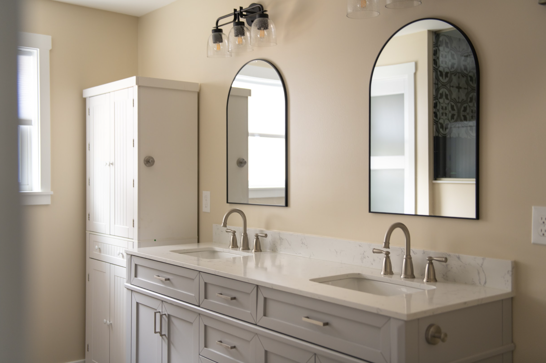 A bathroom vanity with a white marble countertop, two sinks, and two brushed nickel faucets. Above the vanity are two arched mirrors with black frames. To the left, there is a white cabinet with paneled doors. The wall is painted a light beige color, and there are two windows with white trim allowing natural light into the room.