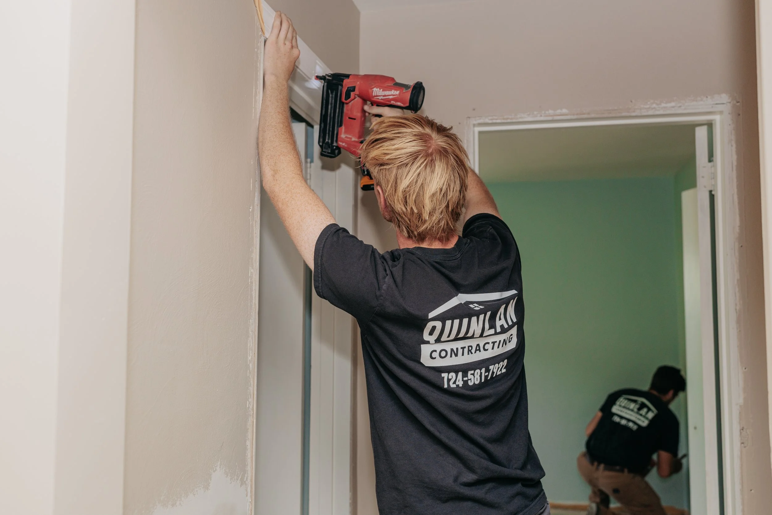 Worker installing or repairing in a home interior wall area using a power nail gun, wearing a black shirt with 'Quinlan Contracting' on the back. Another individual is bent down in the background.