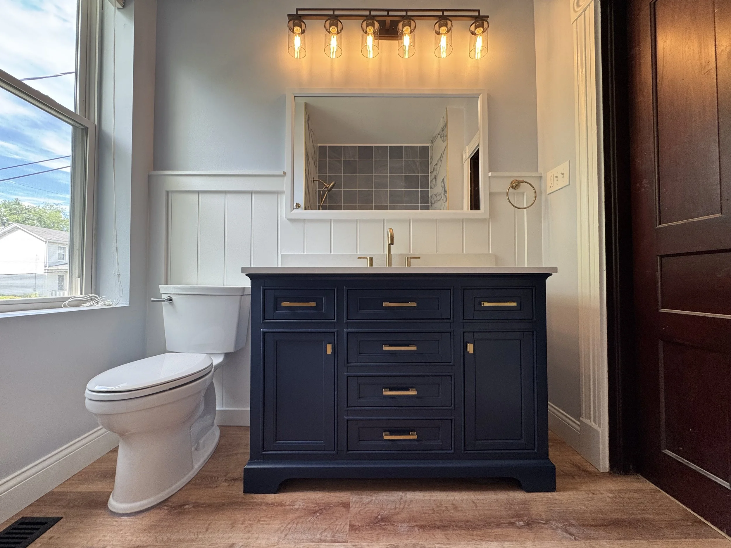 Bathroom interior featuring a white toilet, a navy blue vanity with gold hardware, a large mirror, a window with blinds, and a view of a shower with gray tiles.