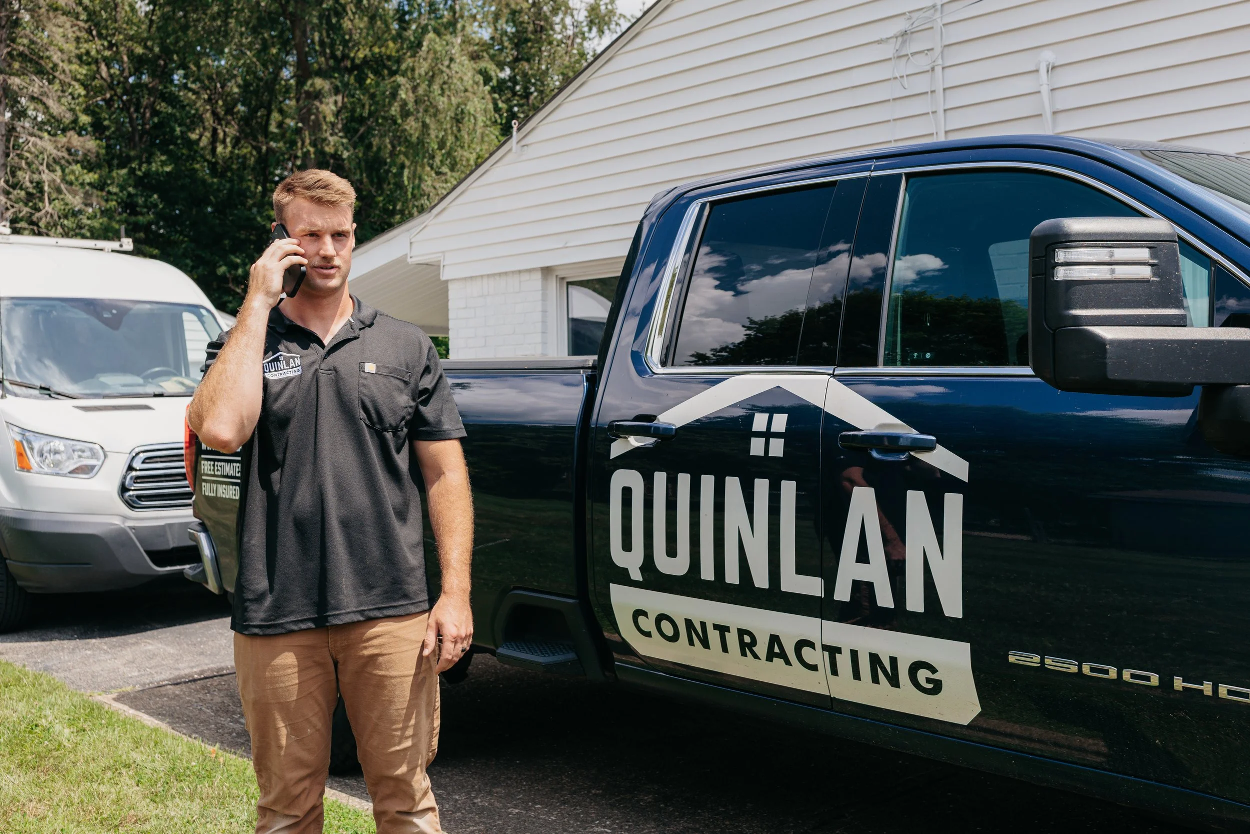 A man in uniform holding a phone to his ear, standing next to a black contracting truck with the company's logo and name, in a residential area with trees and a white house.