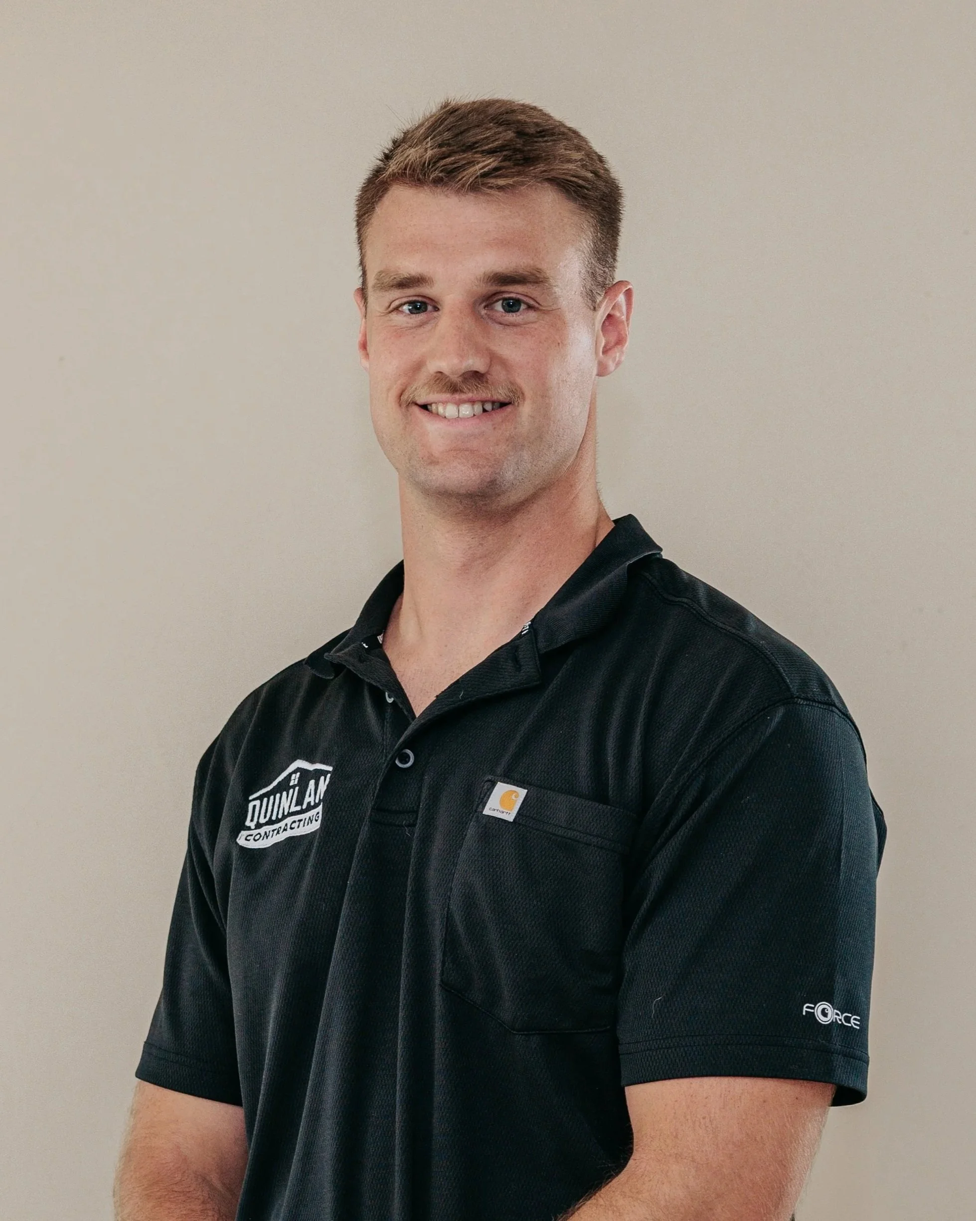 Portrait of a young man with short brown hair, wearing a black Carhartt work shirt with logos, standing against a plain beige wall.