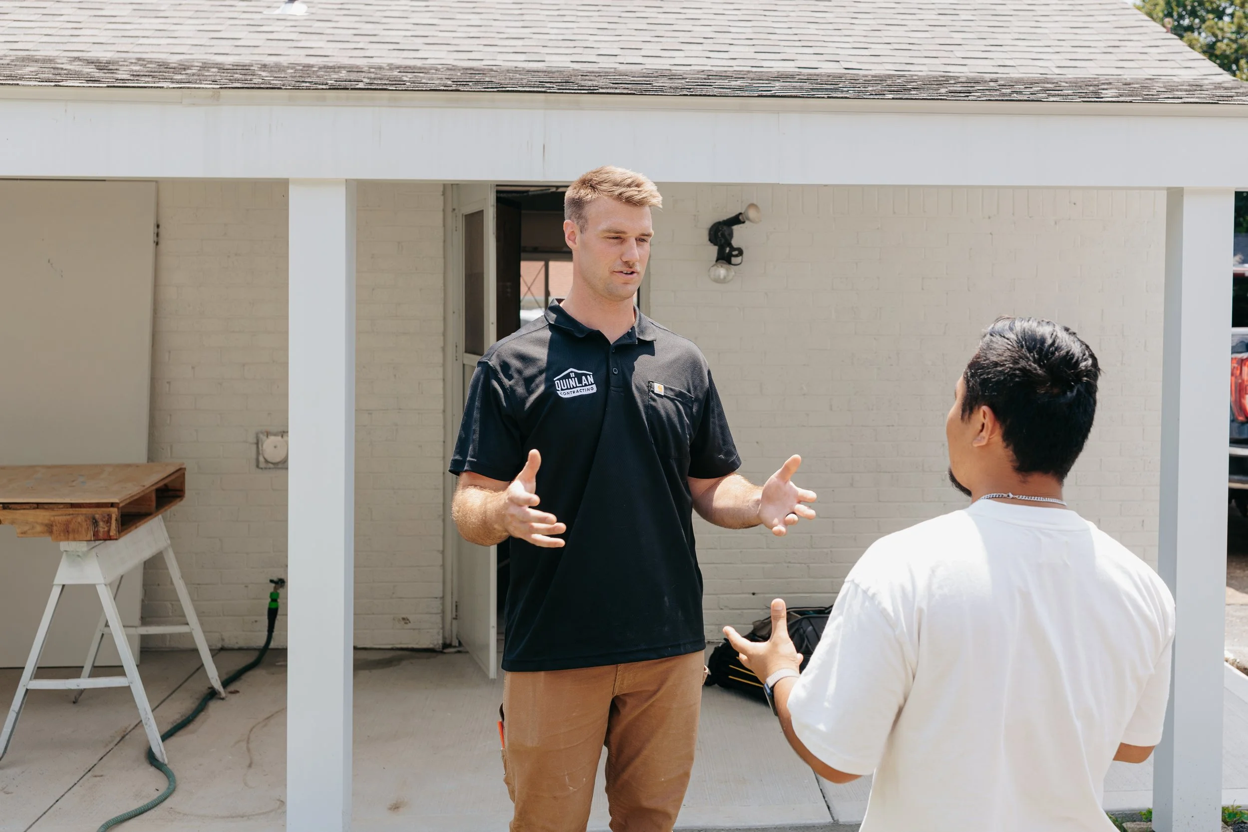 Two men having a discussion outdoors, one wearing a black polo shirt with a logo and the other wearing a white shirt, in front of a white brick building with a partially constructed frame.