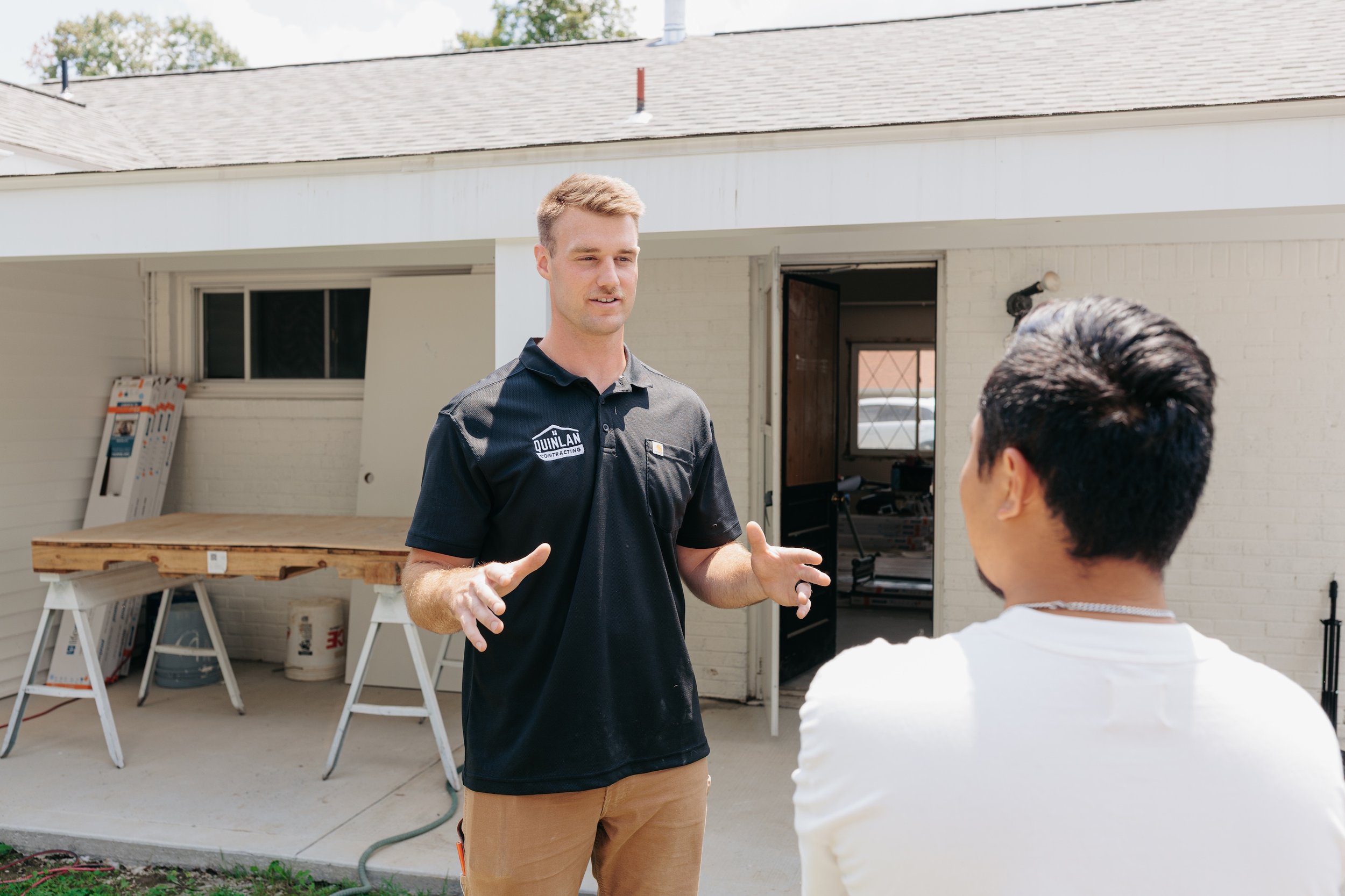 A man wearing a black shirt with 'Quinlan Contracting' on it is talking to another person outside a house. The house's exterior is white with an open door and some construction materials.