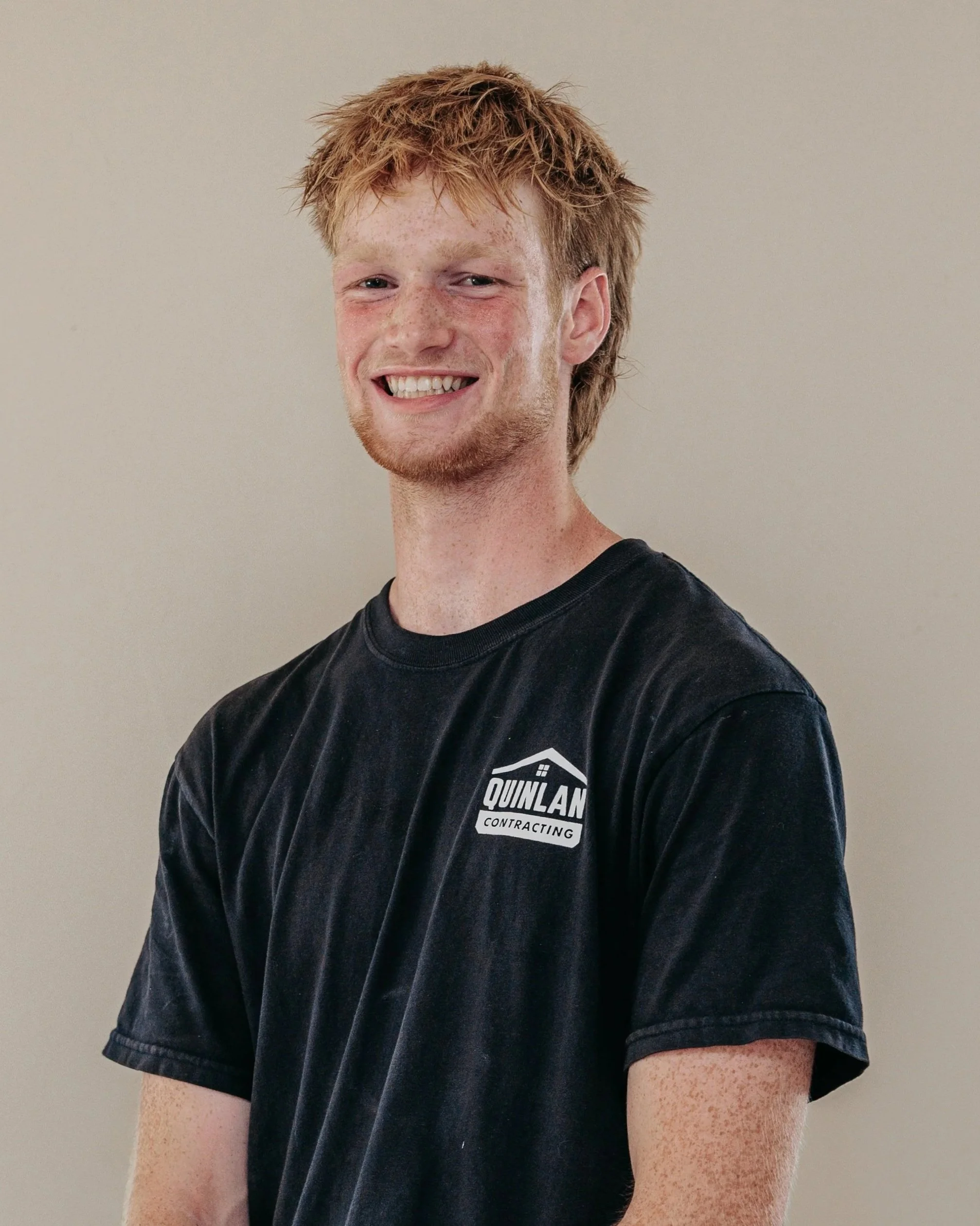 Close-up of a young man with red hair, freckles, and a beard, smiling and wearing a black T-shirt with a logo that reads "Qunlan Contracting" against a plain background.