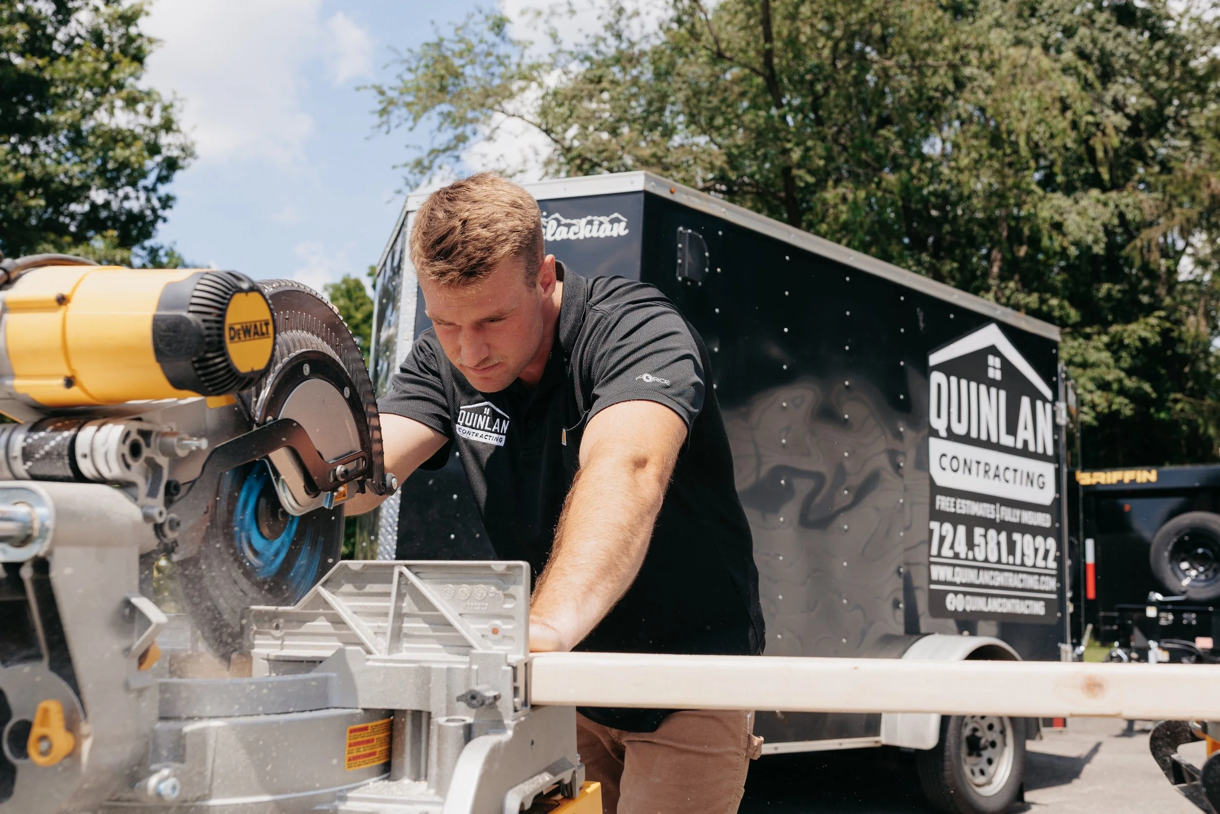 A man working with a circular saw outdoors near a black vehicle trailer for Quinlan Contracting.