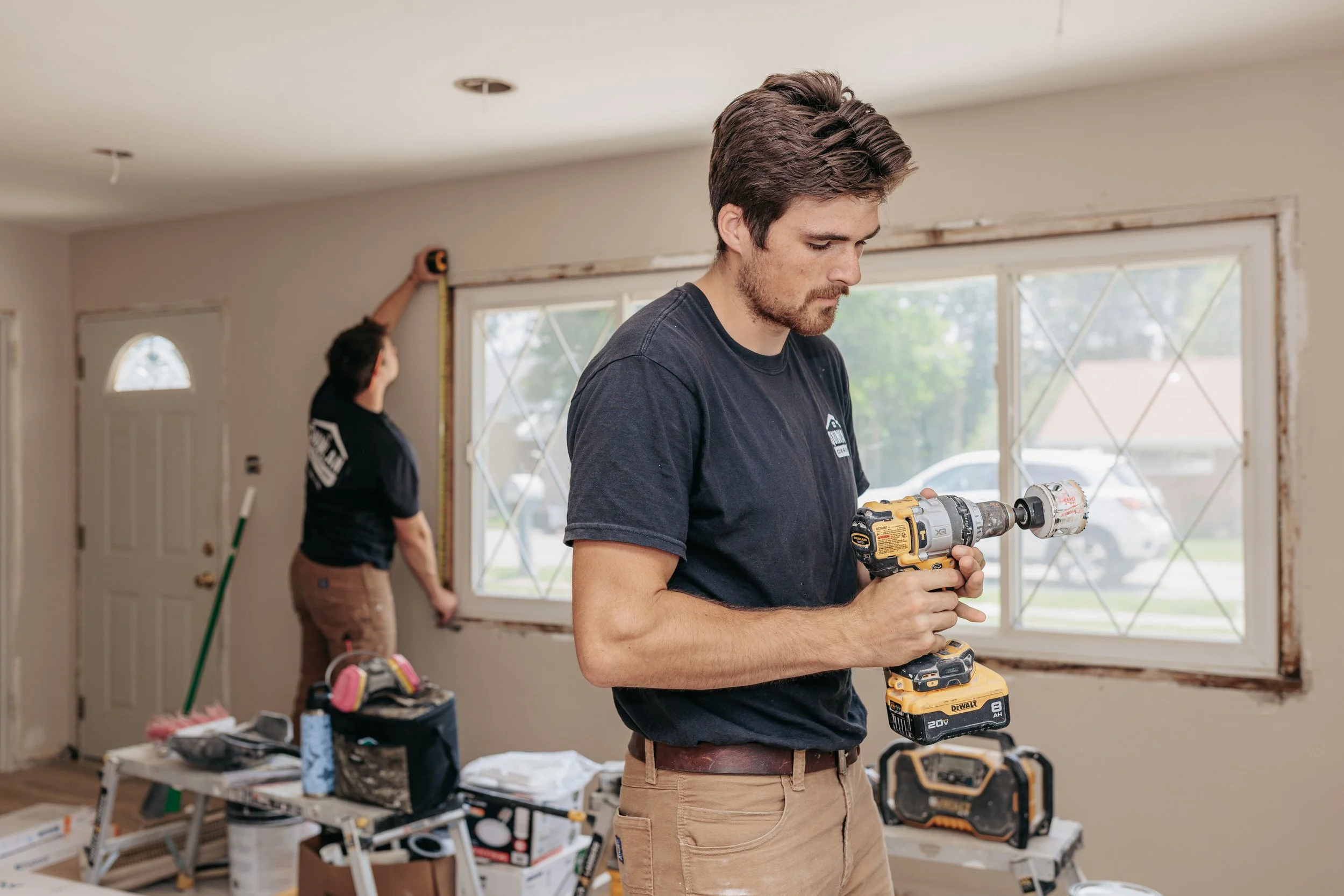 A young man is working with a power drill in a room under renovation. Behind him, another person measures a window frame. The room contains construction tools, supplies, and furniture covered for protection.