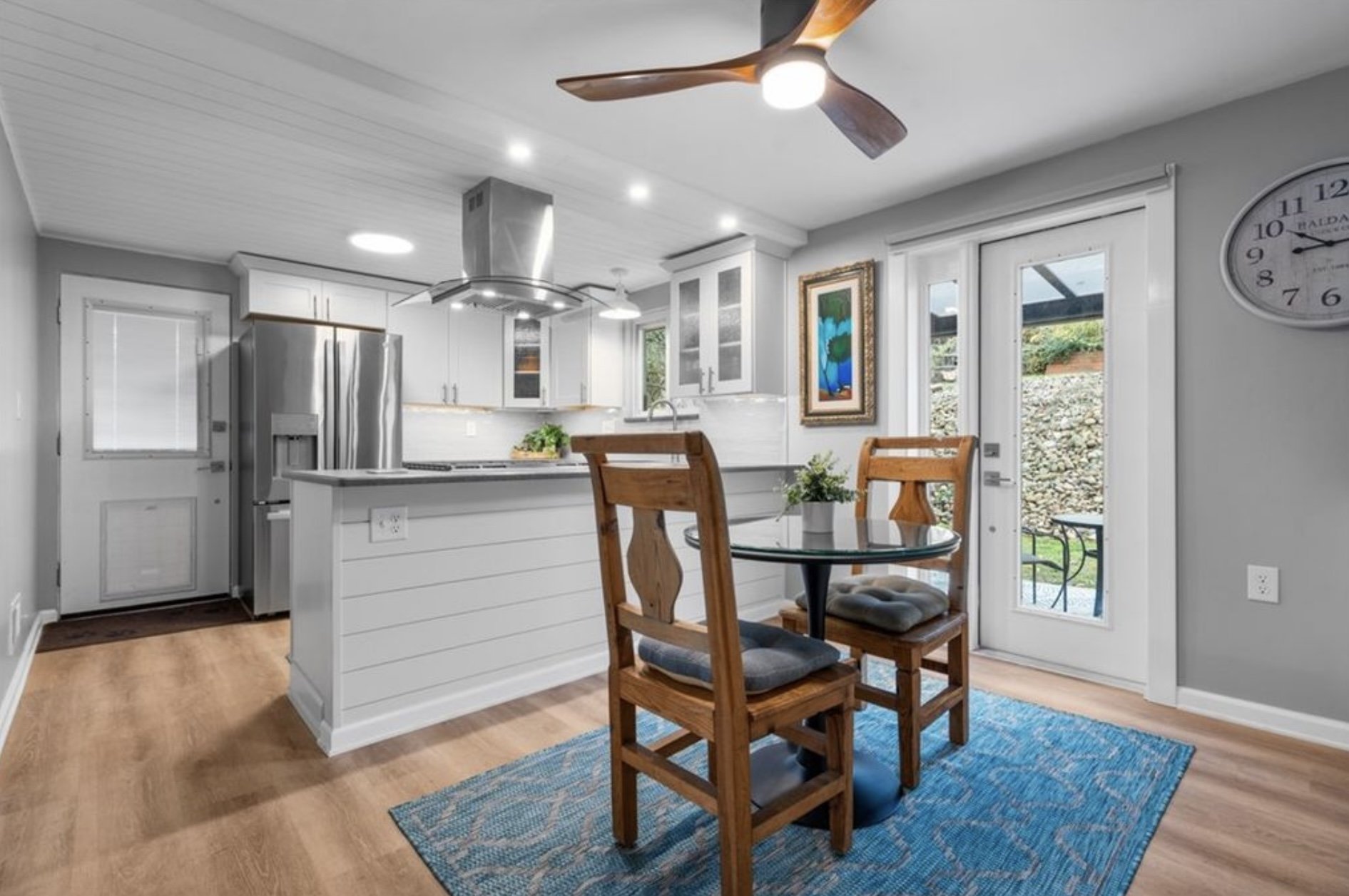 Interior view of a modern kitchen with white cabinets, stainless steel refrigerator, and a small dining area with a round glass table and wooden chairs.
