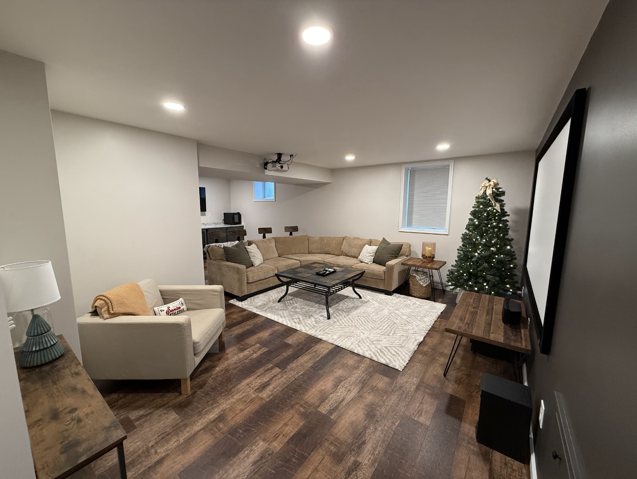 A cozy basement living room decorated for Christmas with a small Christmas tree, a sectional sofa, an armchair, a coffee table, and a projector screen on the wall.