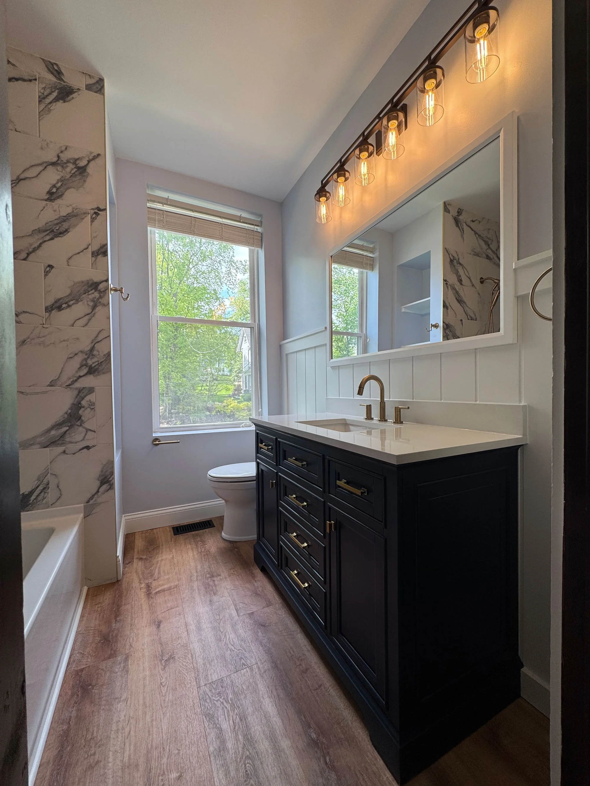A bathroom with large windows, a dark blue vanity with numerous drawers, a white countertop with a gold faucet, a mirror above the vanity, and a light fixture with six Edison-style bulbs. The bathroom has wood flooring and partial marble tile walls.