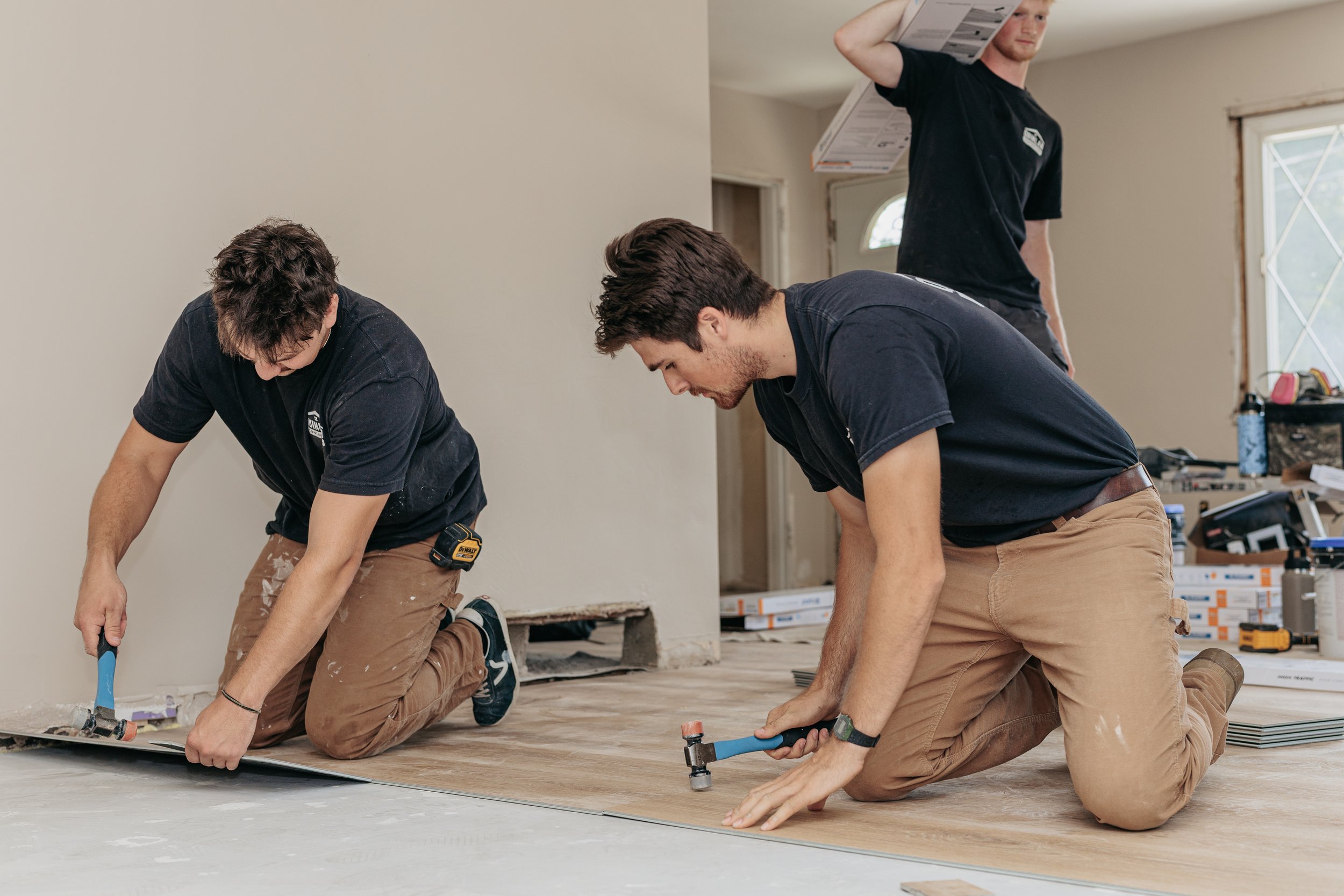 Three men kneeling on the floor installing a wooden floor, working together with tools, inside a house with unfinished walls and a window in the background.