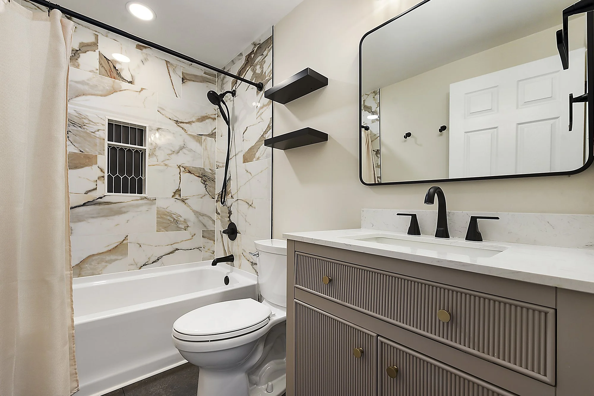 Modern bathroom with marble-tiled walk-in shower, black shower head, three black floating shelves, large mirror, beige vanity with black fixtures, and white door.