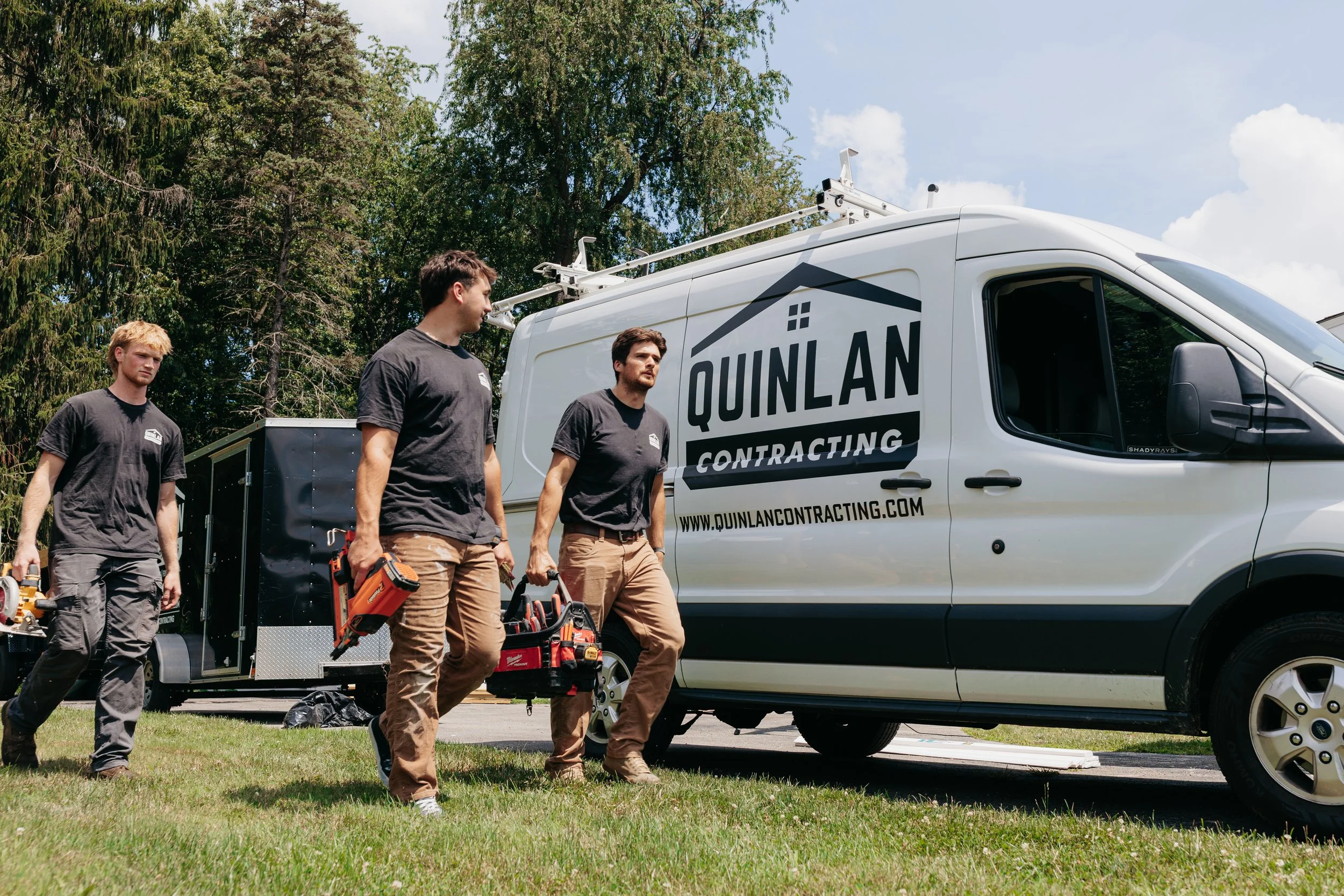 Three construction workers walking away from a white van with 'Quinlan Contracting' branding, carrying tools outdoors on a sunny day with trees in the background.