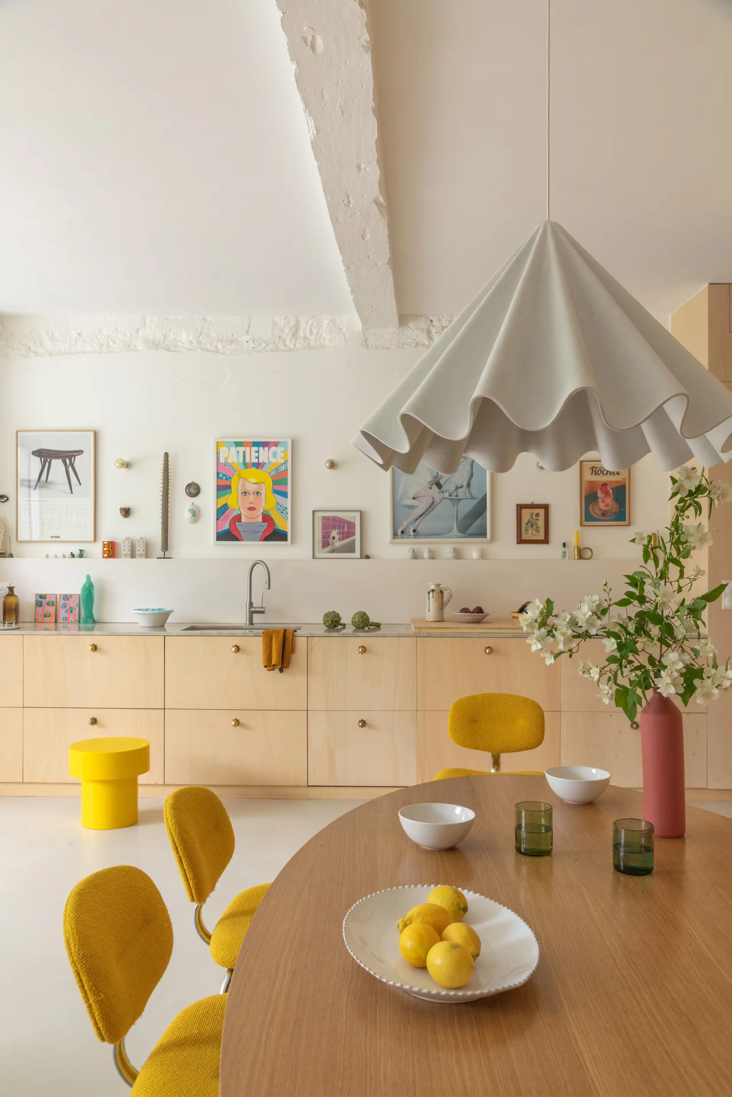 Une cuisine moderne avec un plan de travail en bois clair, des armoires blanches, des chaises jaunes, un vase rose avec des fleurs blanches, un plafond avec un plafond en pente et un luminaire en forme d'ombrelle. Il y a un tableau coloré avec la phrase 'Patience' et divers objets décoratifs.