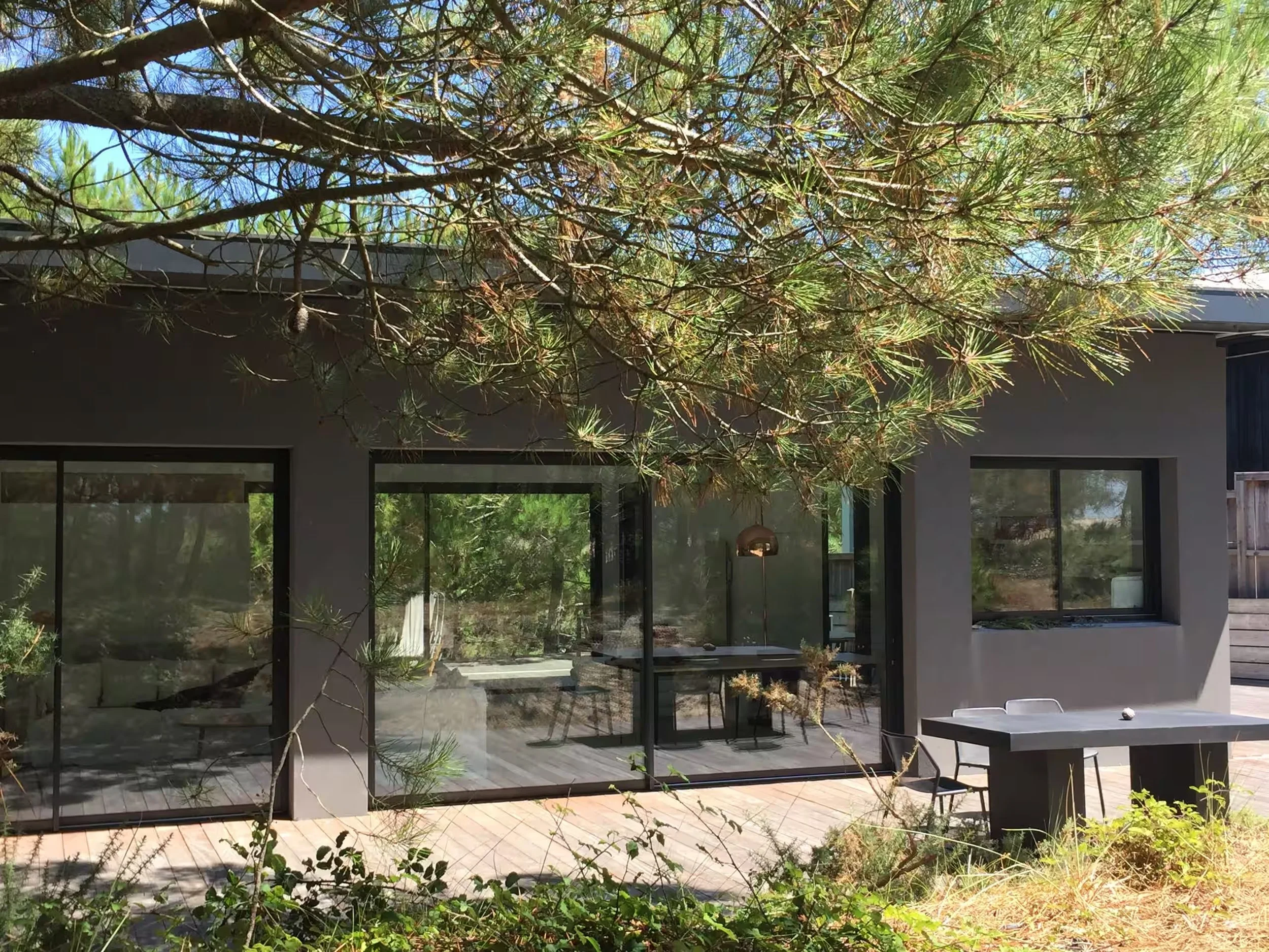 Une maison moderne avec de grandes fenêtres en verre et un toit plat est visible à travers des branches d'arbres verts. Il y a une terrasse en bois avec une table et des chaises en plein air. Le ciel est clair et ensoleillé.