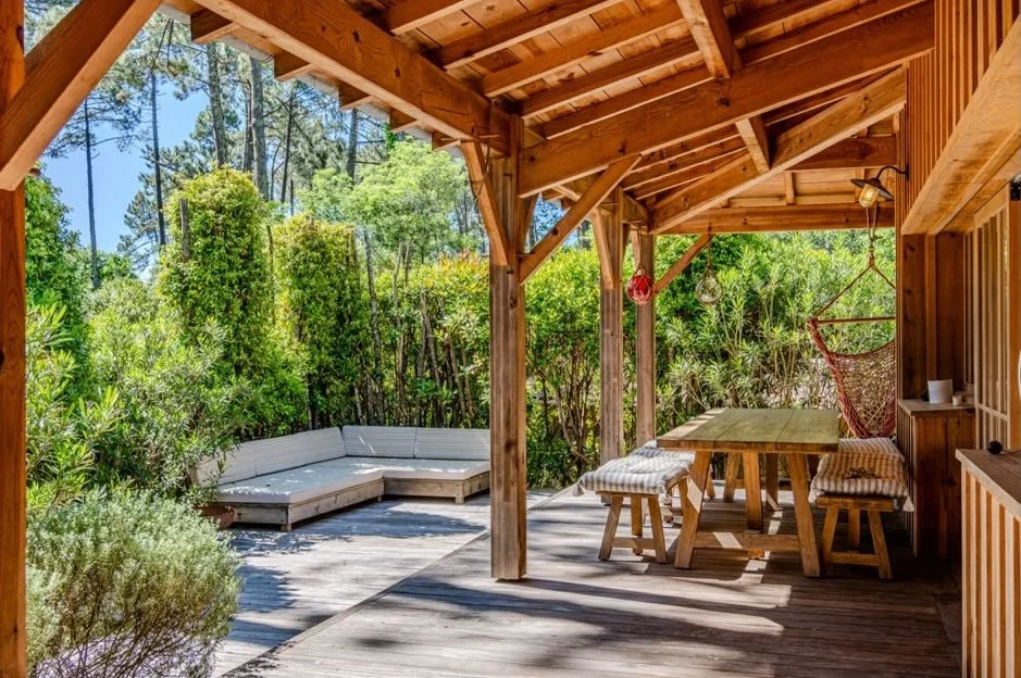 Une terrasse en bois avec mobilier, entourée de verdure et sous un toit en bois, avec un canapé en coin blanc, une table en bois et des coussins.