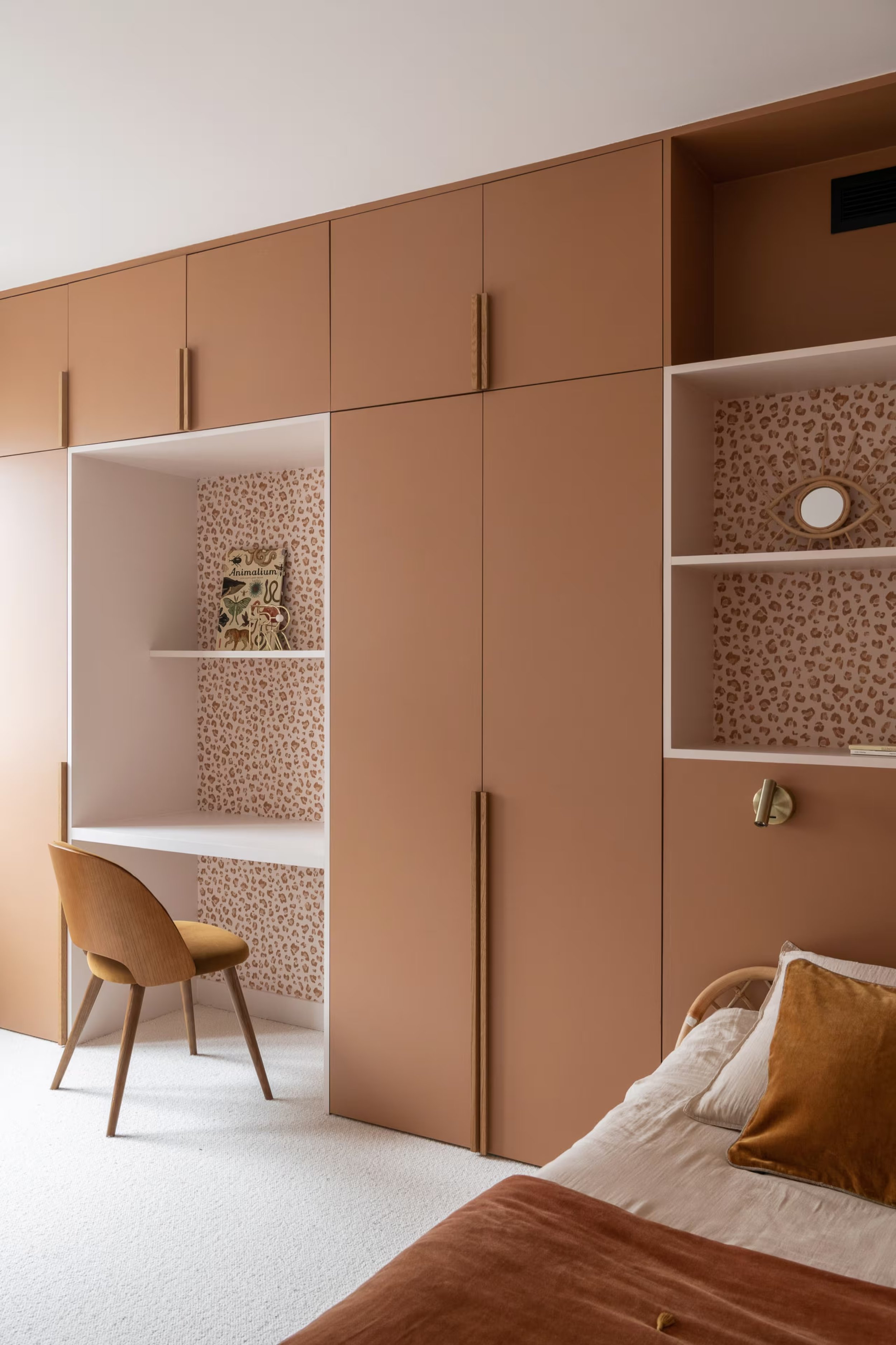 Intérieur d'une chambre avec un mur en placard en bois clair, une étagère avec décoration murale, un lit avec des oreillers en coussin marron et beige, une chaise en bois clair.
