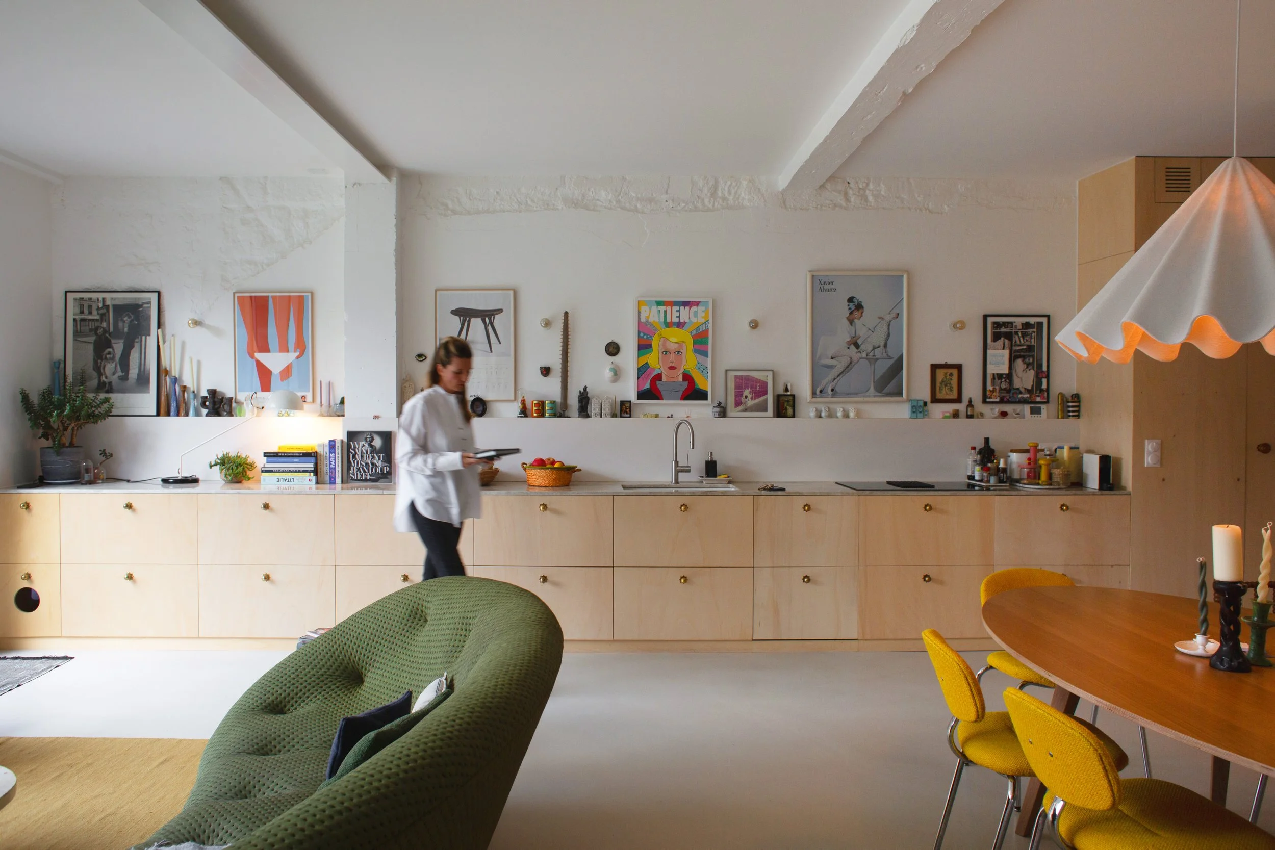Intérieur d'une cuisine moderne avec un mur blanc décoré de tableaux, un comptoir en bois clair, un évier, et une personne portant un tablier blanc. Une table ronde en bois avec des chaises jaunes est visible à droite, et un canapé vert est en premier plan.