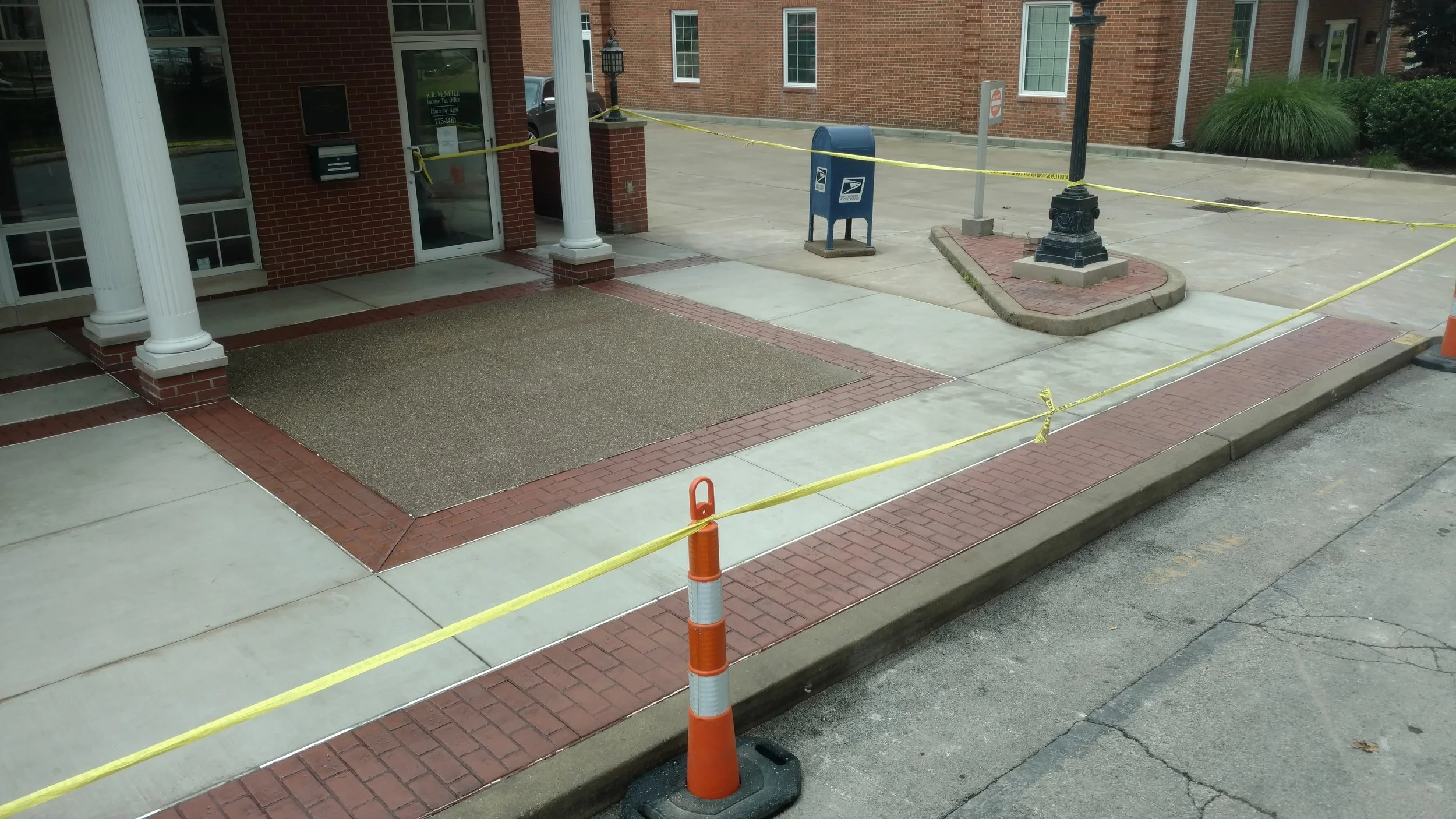 Sidewalk outside a brick building enclosed by yellow caution tape, with a mailbox, streetlamp, and brick-paved areas.
