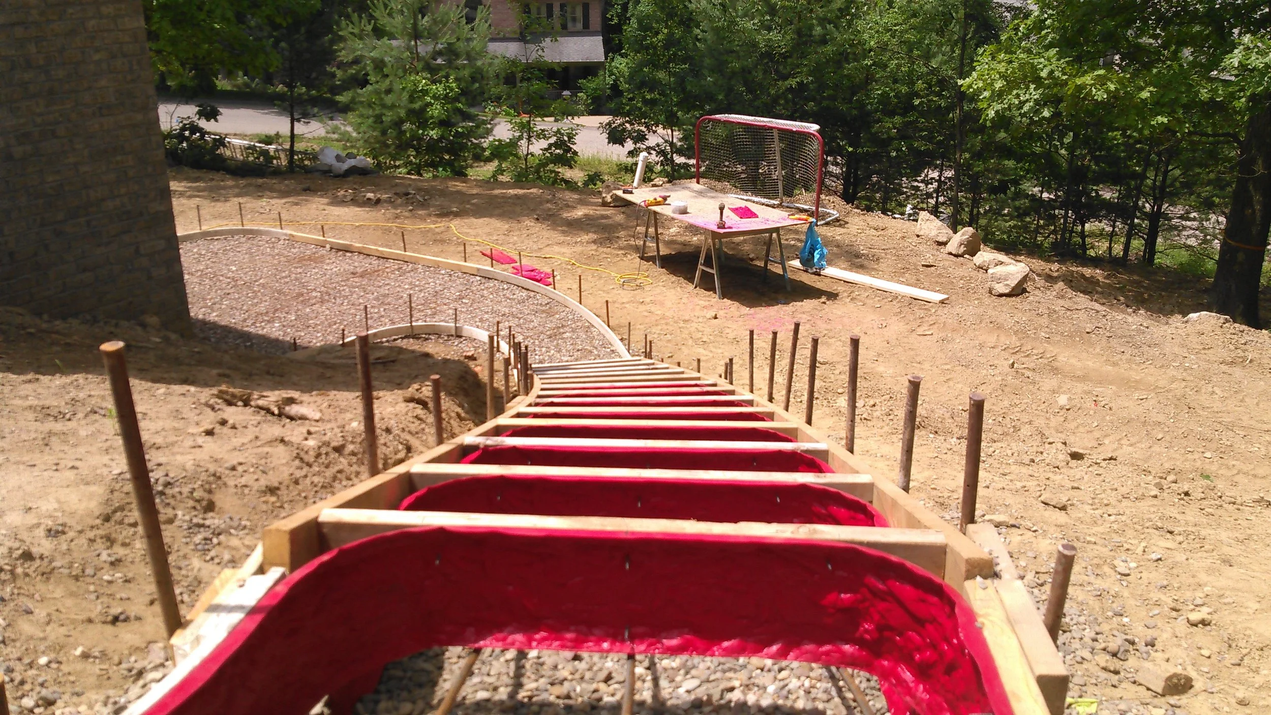 Outdoor construction site with a wooden staircase leading down a dirt slope, a gravel area, and a work table with tools, surrounded by green trees and a residential building in the background.
