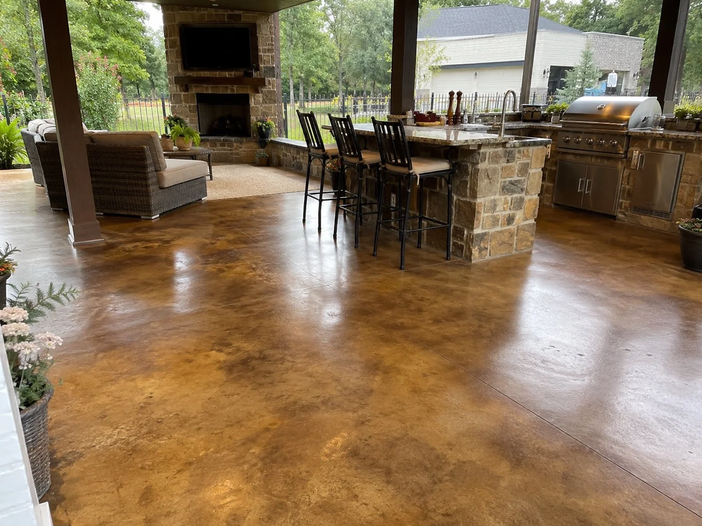 Covered outdoor patio with a polished concrete floor, an outdoor kitchen with a stone countertop, a grill, and barstools, a cozy seating area with a wicker armchair and a rug, a brick fireplace with a mounted TV, and lush green trees visible through screened windows.