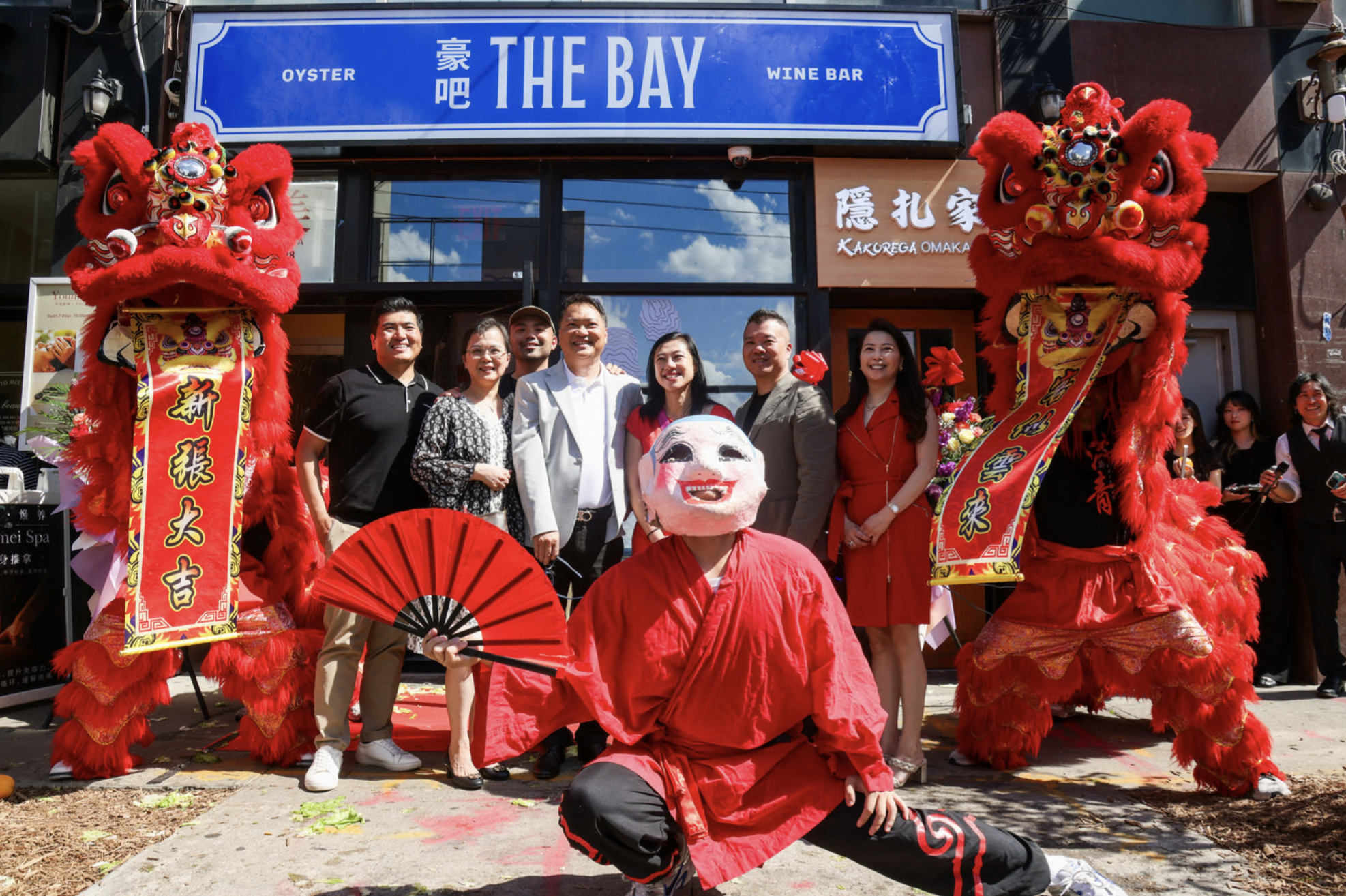 A group celebrating outside a restaurant called 'The Bay' with Chinese New Year decorations, including lion dance costumes, a paper mask, and people in festive attire.
