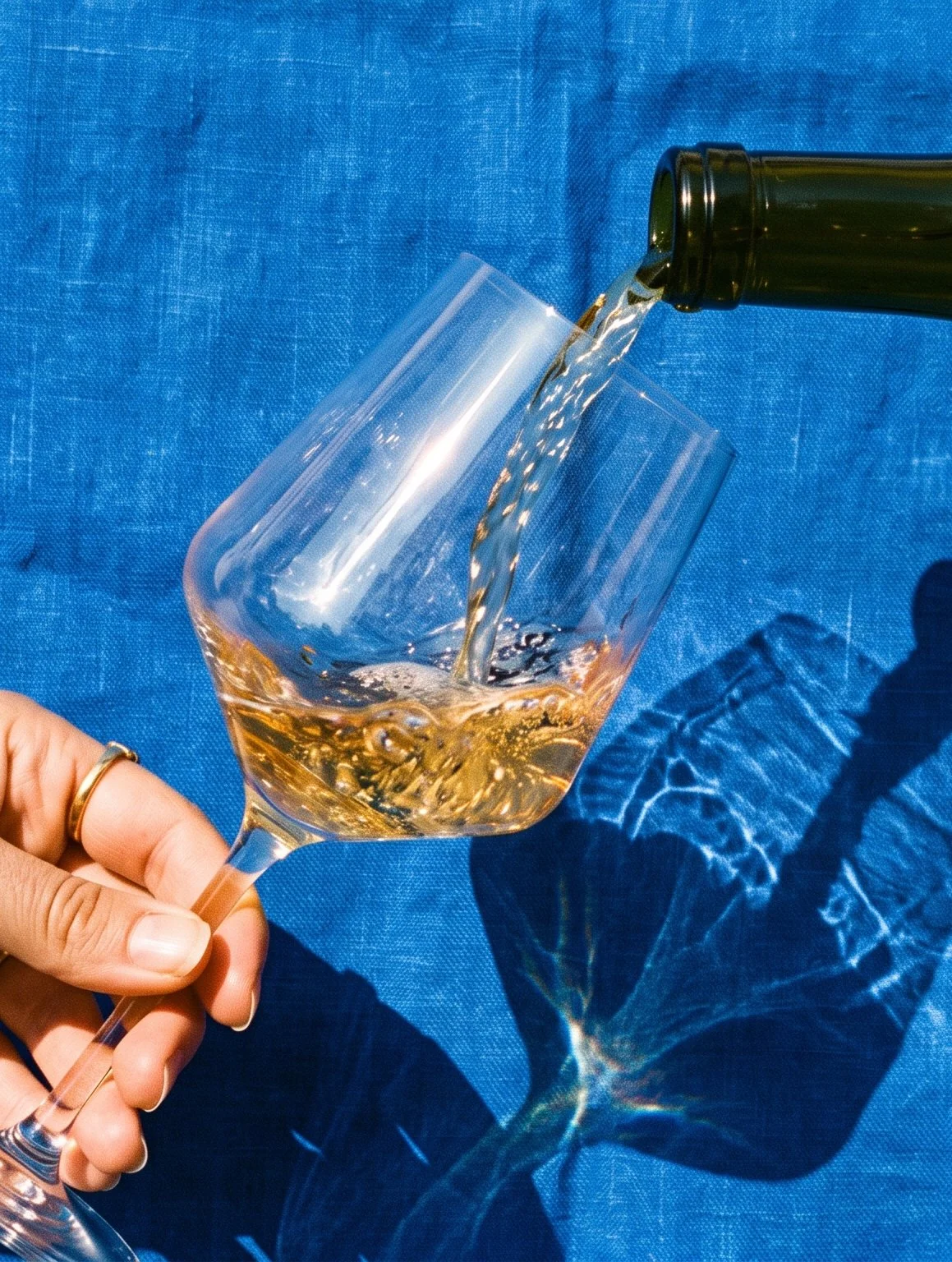 A hand holding a wine glass as rosé wine is being poured from a bottle, with a blue textured background and shadows on the surface.