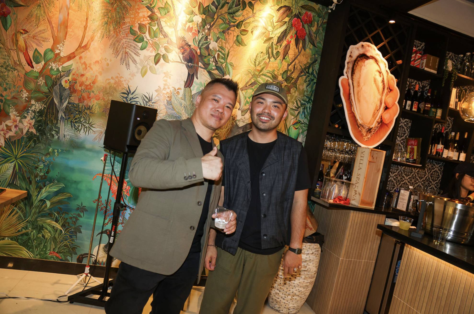 Two men standing in a colorful, tropical-themed restaurant with a mural of birds and plants on the wall behind them. One man is wearing a gray blazer and the other is in a black shirt and cap. The man in the gray blazer is holding a drink and giving a thumbs-up.