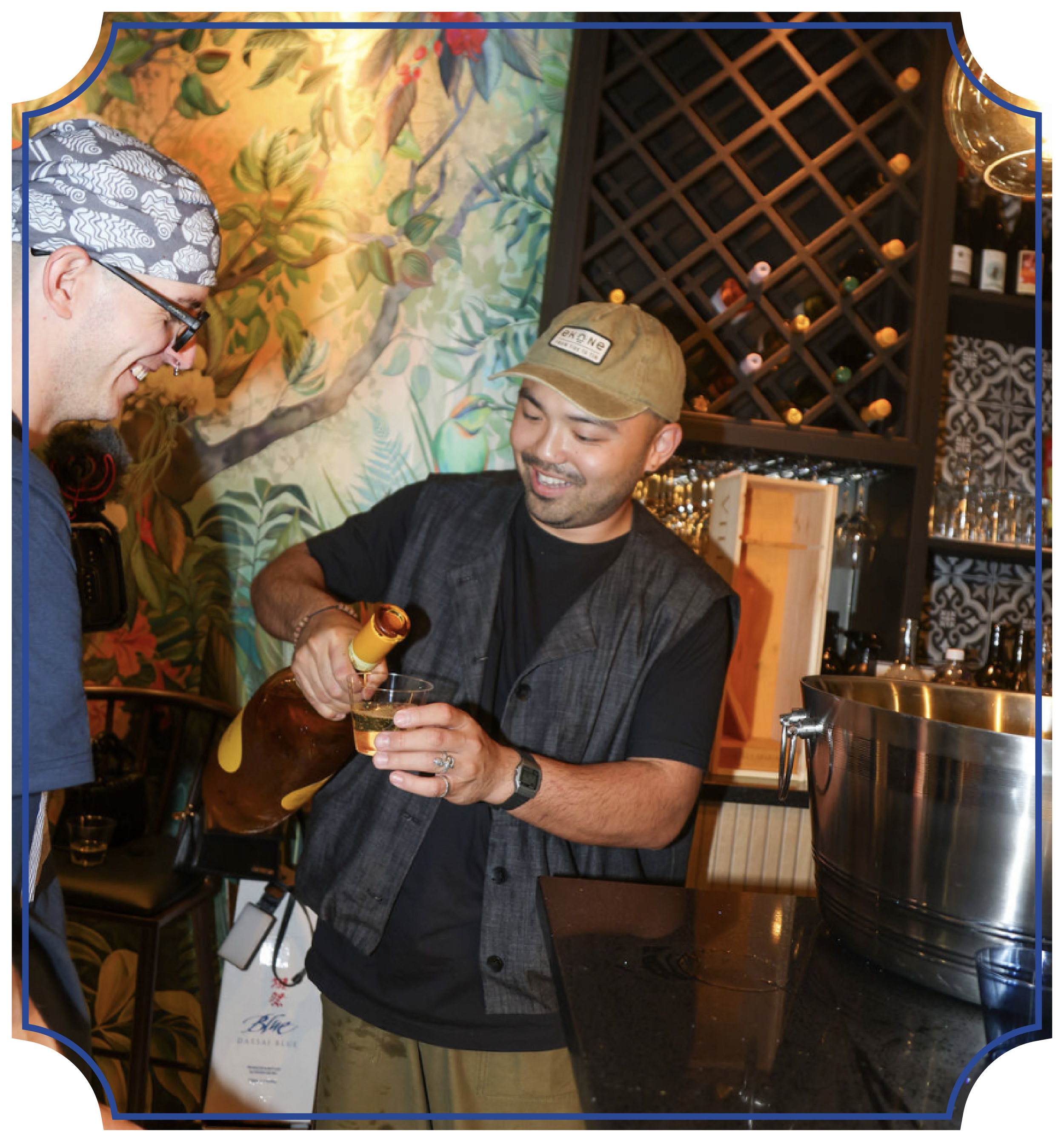 Two men smiling, one pouring a drink into a glass, in a restaurant or bar setting with wine bottles and a decorative wall in the background.