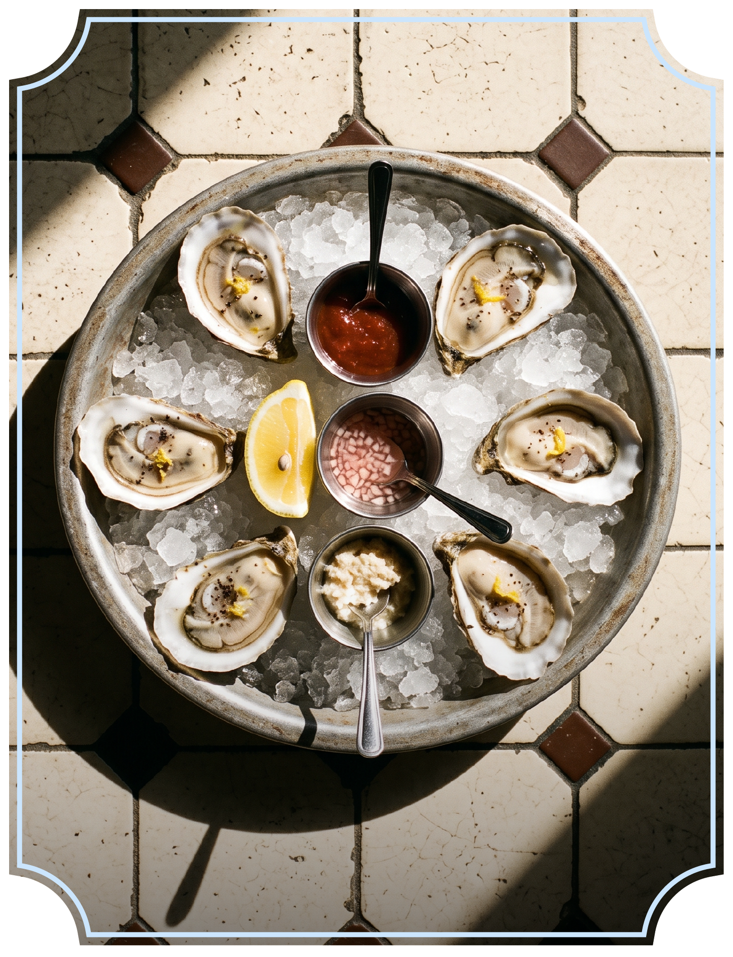 A round metal tray filled with crushed ice, holding six raw oysters on the half shell, each garnished with lemon wedge, with small containers of cocktail sauce, tarama, and horseradish, on a tiled floor with sunlight casting shadows.
